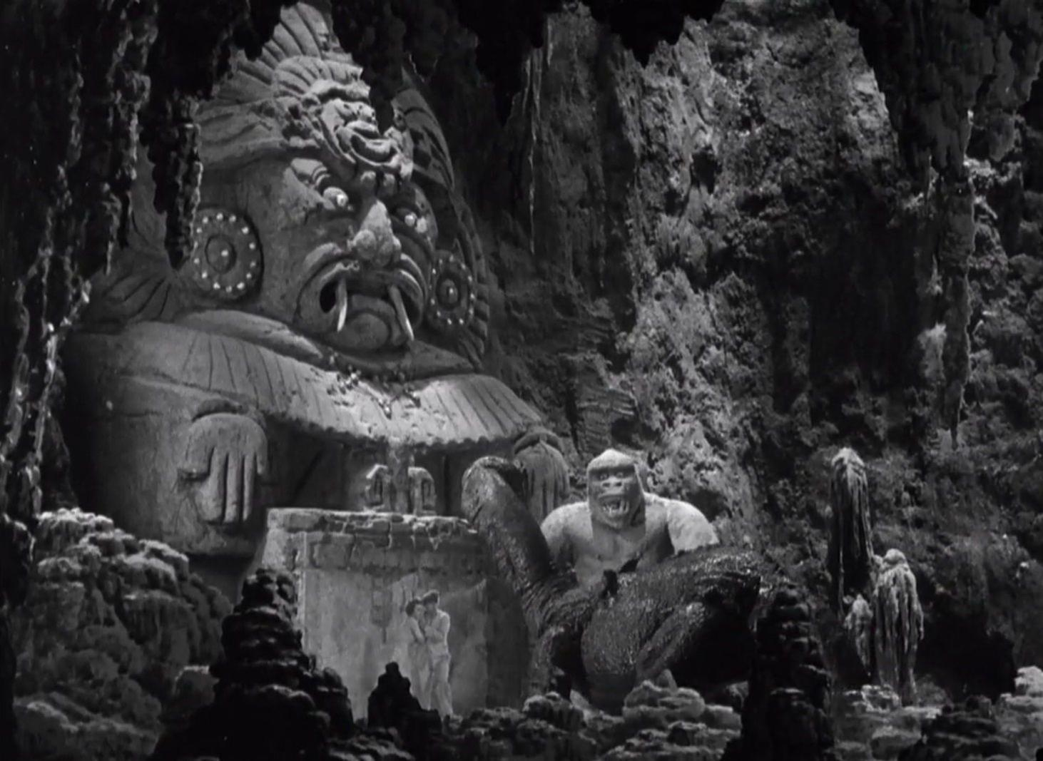 Ein Gorilla und ein Dinosaurier stehen vor einer riesigen Steinskulptur in einer Höhle.