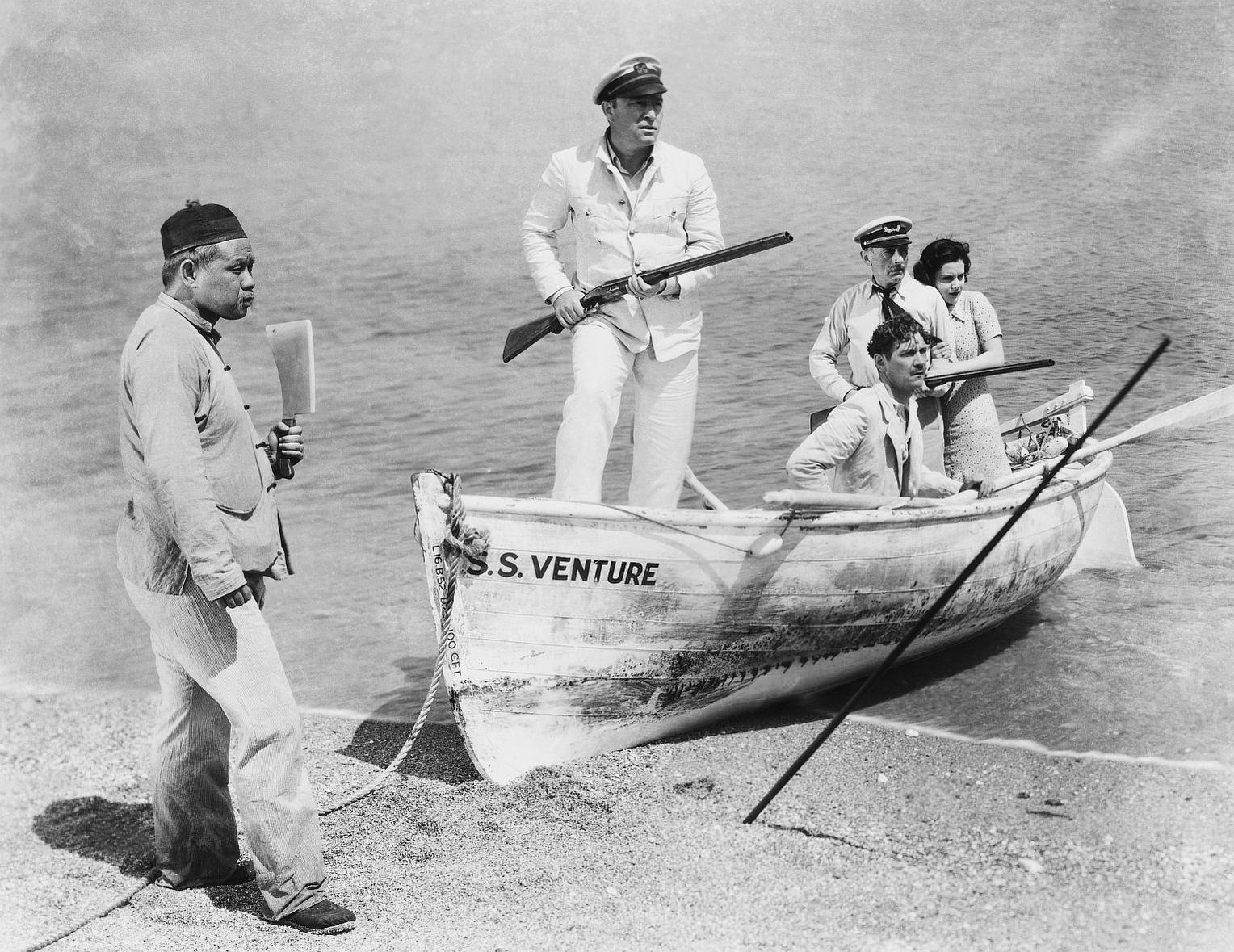 Ein Mann mit einem Gewehr steht in einem Boot. Andere stehen im Boot und am Strand.
