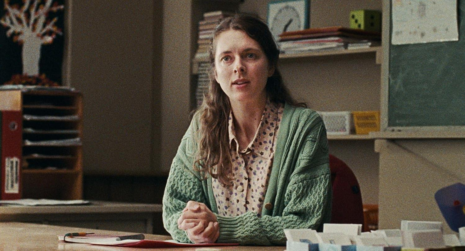 Eine Frau sitzt an einem Schreibtisch in einem Klassenzimmer und trägt eine grüne Strickjacke.