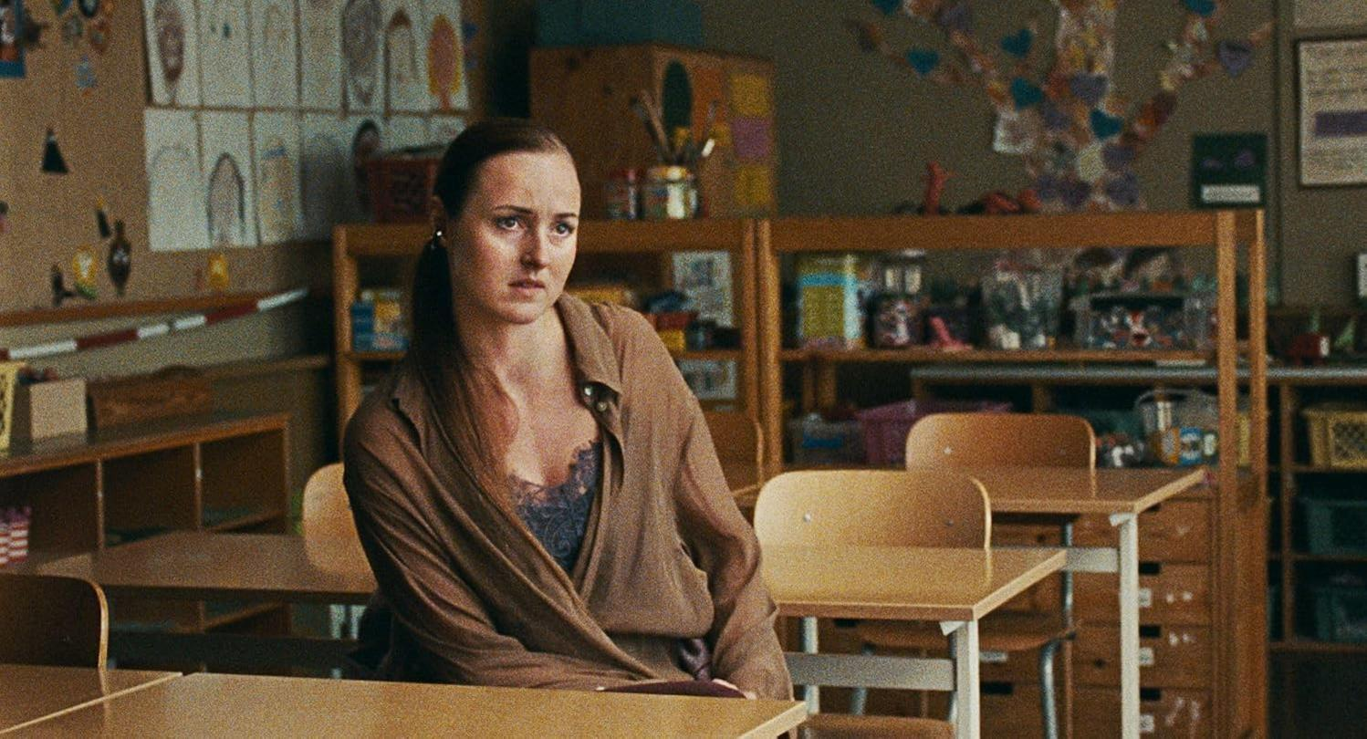 Eine Frau sitzt in einem Klassenzimmer mit Holzmöbeln und Kinderzeichnungen.