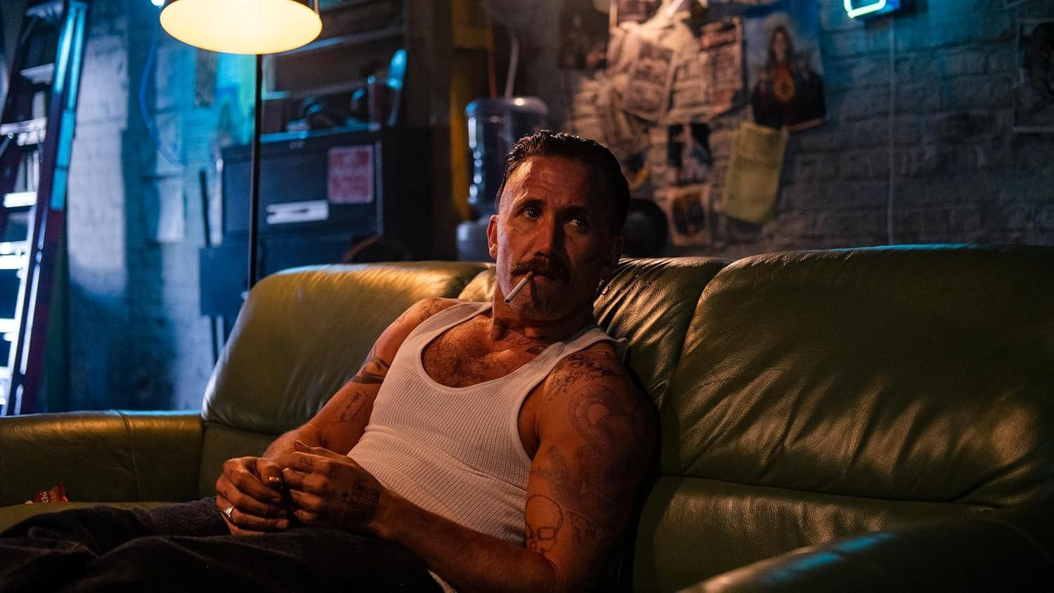 Ein Mann mit Tattoos sitzt auf einem Ledersofa und raucht eine Zigarette.
