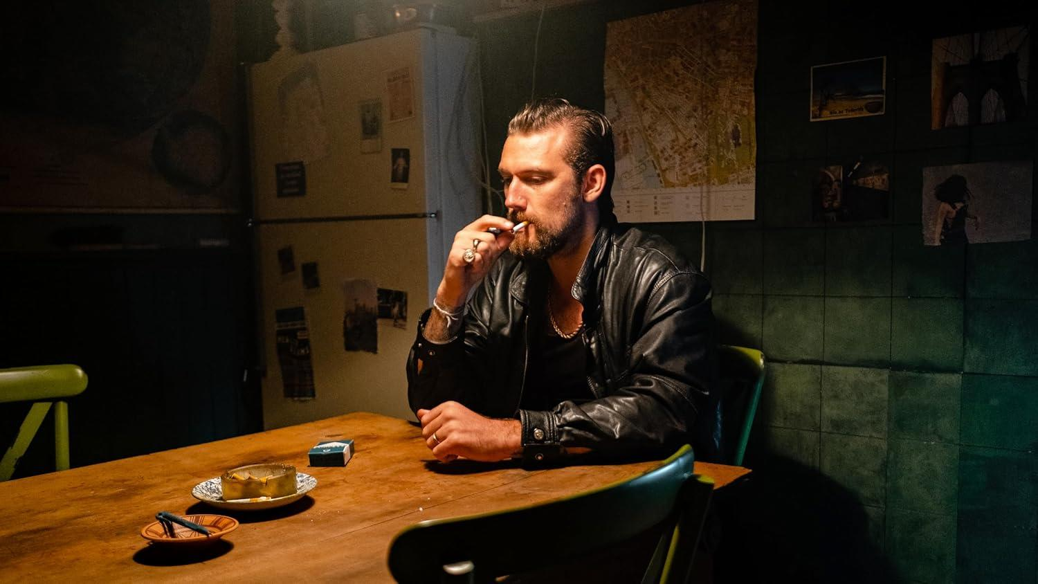 Mann in Lederjacke raucht Zigarette an einem Holztisch mit Aschenbecher.