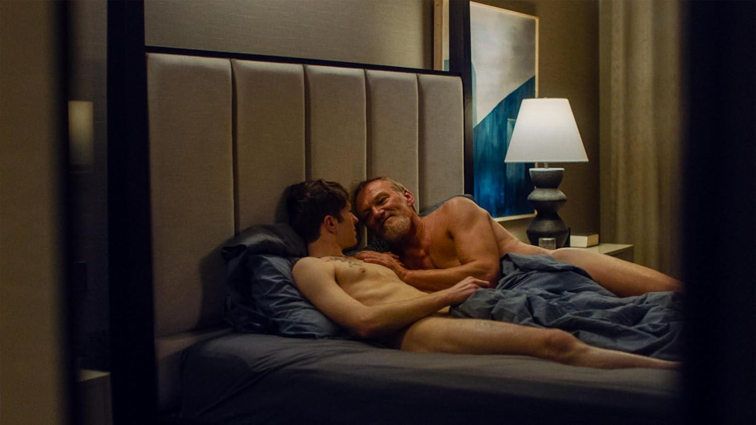 Zwei nackte Männer liegen im Bett und schauen sich an. Einer hat eine Glatze und Bart.