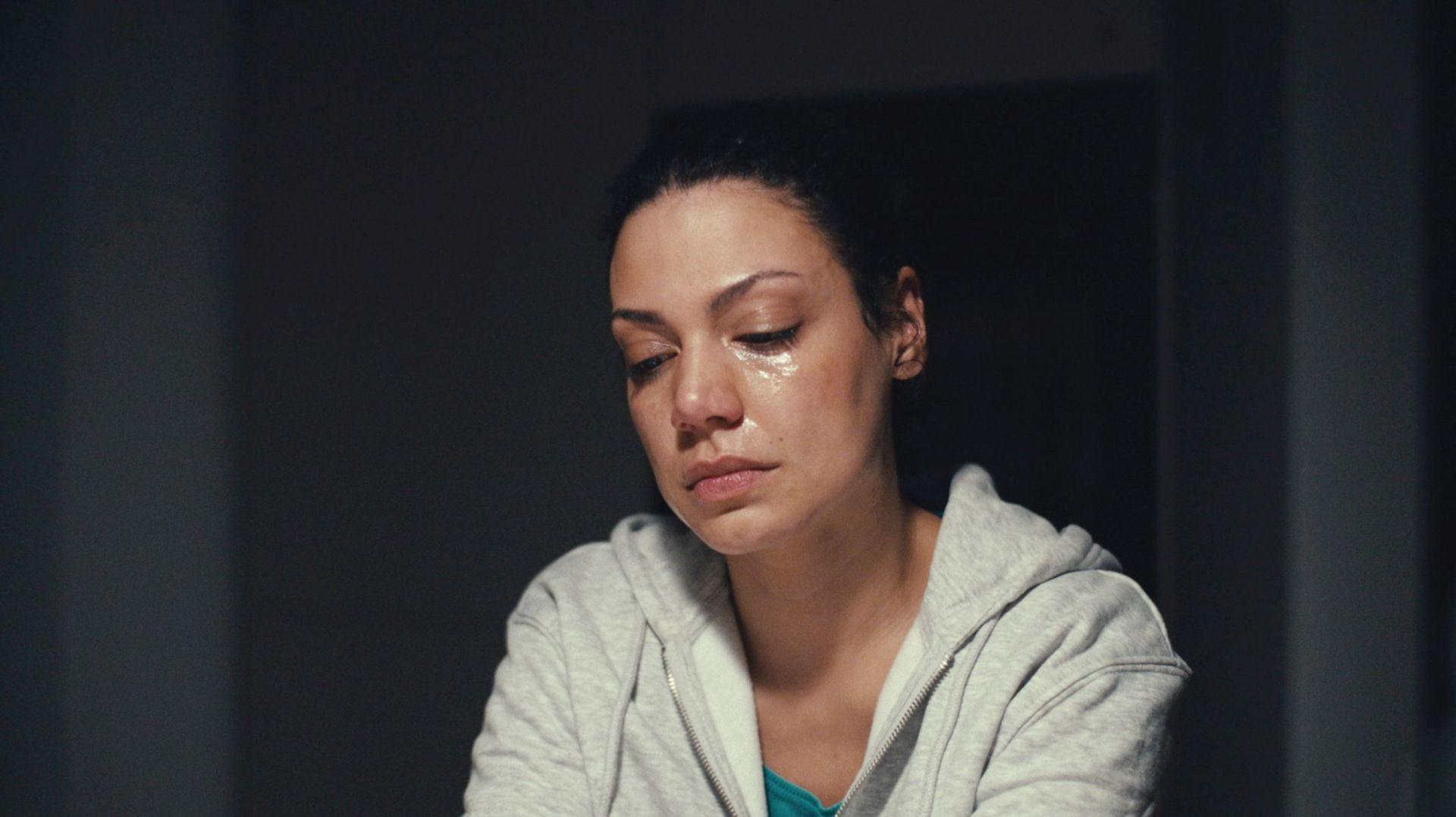 Eine Frau mit tränenfeuchten Augen blickt nach unten, trägt einen grauen Hoodie.