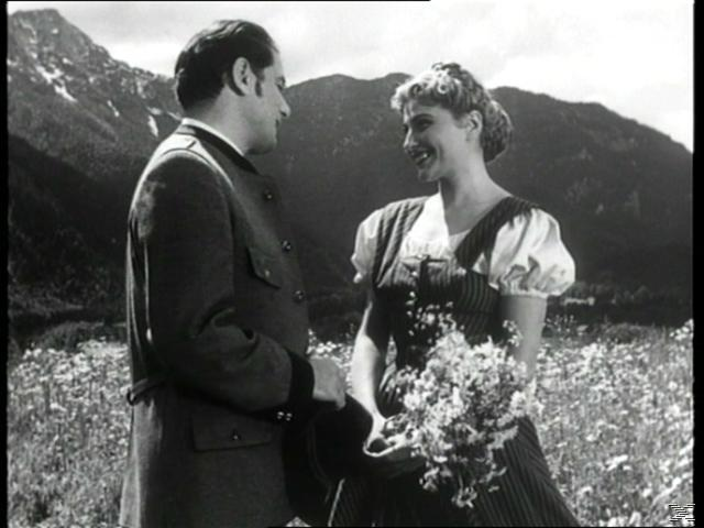 Mann und Frau stehen in einer Blumenwiese vor Bergen. Sie halten Blumen.