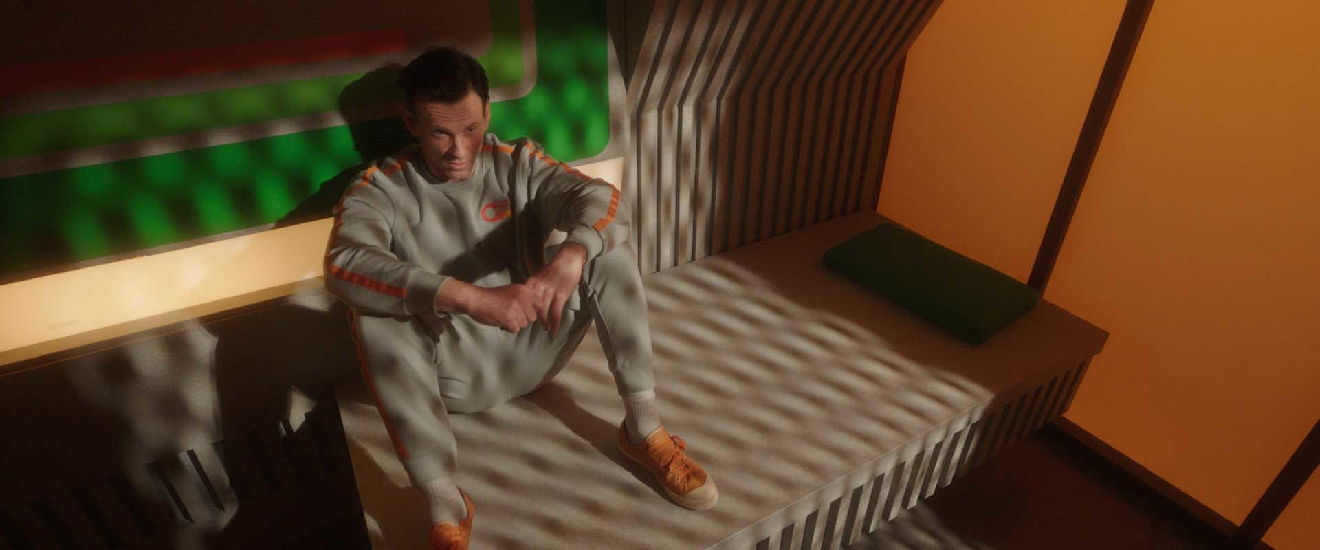 Ein Mann in einem grauen Jogginganzug sitzt auf einer Bank in einem orange-grünen Raum.