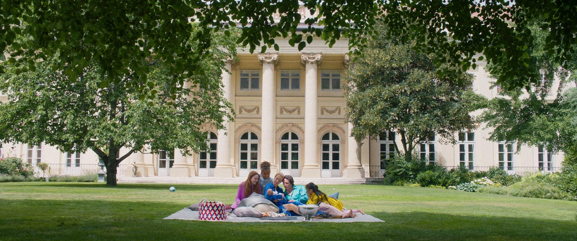 Familie picknickt im Garten vor einem Gebäude mit Säulen.