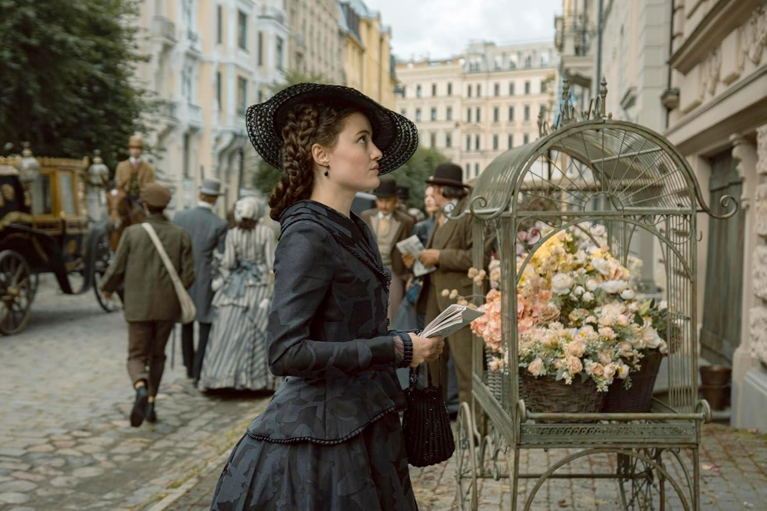 Eine Frau in einem dunkelblauen Kleid und Hut steht vor einem Blumenwagen in einer Stadt.