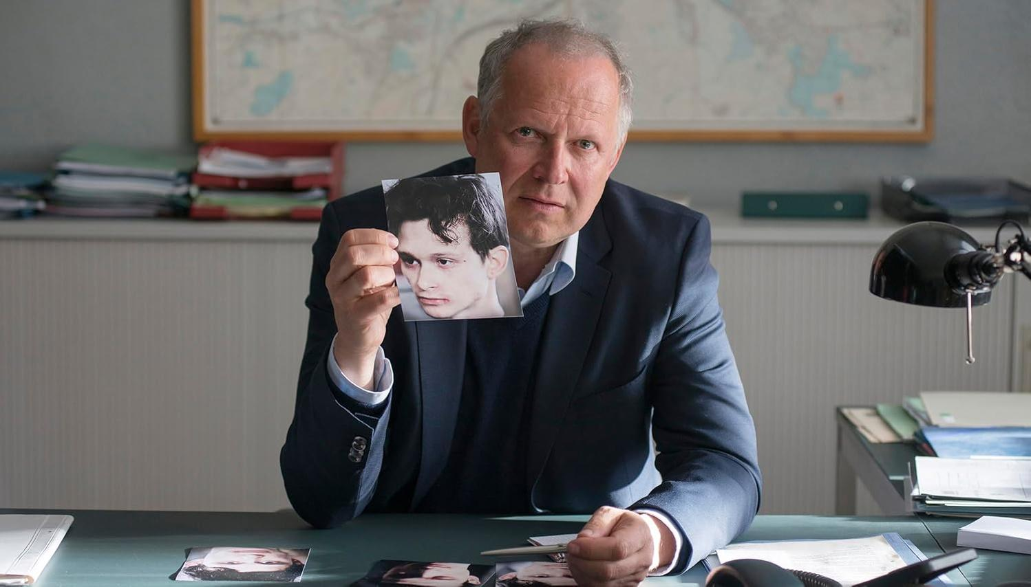 Ein Mann hält ein Foto eines jungen Mannes. Mehr Fotos liegen auf dem Tisch.