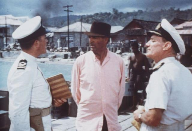 Drei Männer in einem Hafen. Zwei tragen weiße Uniformen, einer ein rosa Hemd.