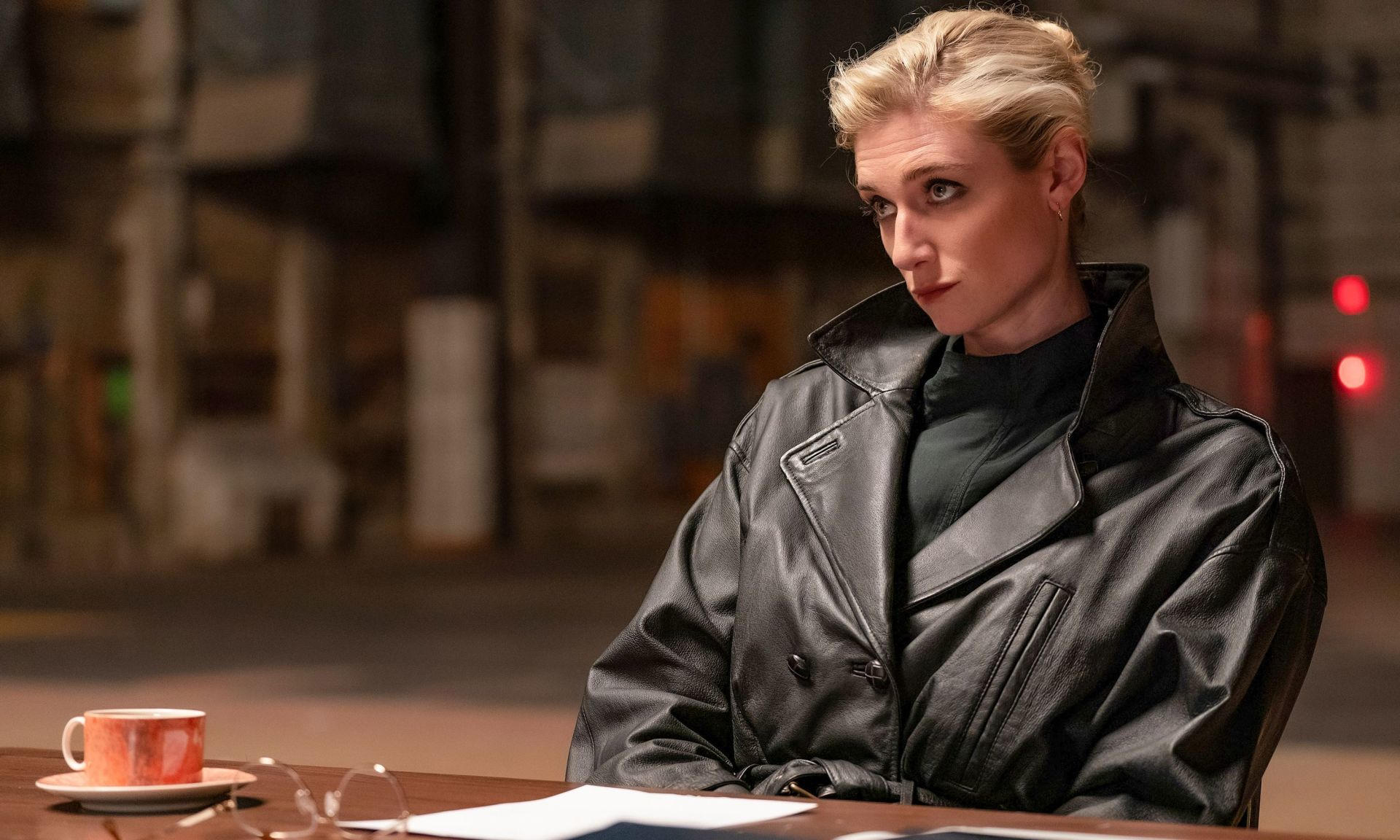 Eine Frau mit blonden Haaren trägt eine schwarze Lederjacke und sitzt an einem Tisch mit einer Tasse Kaffee.