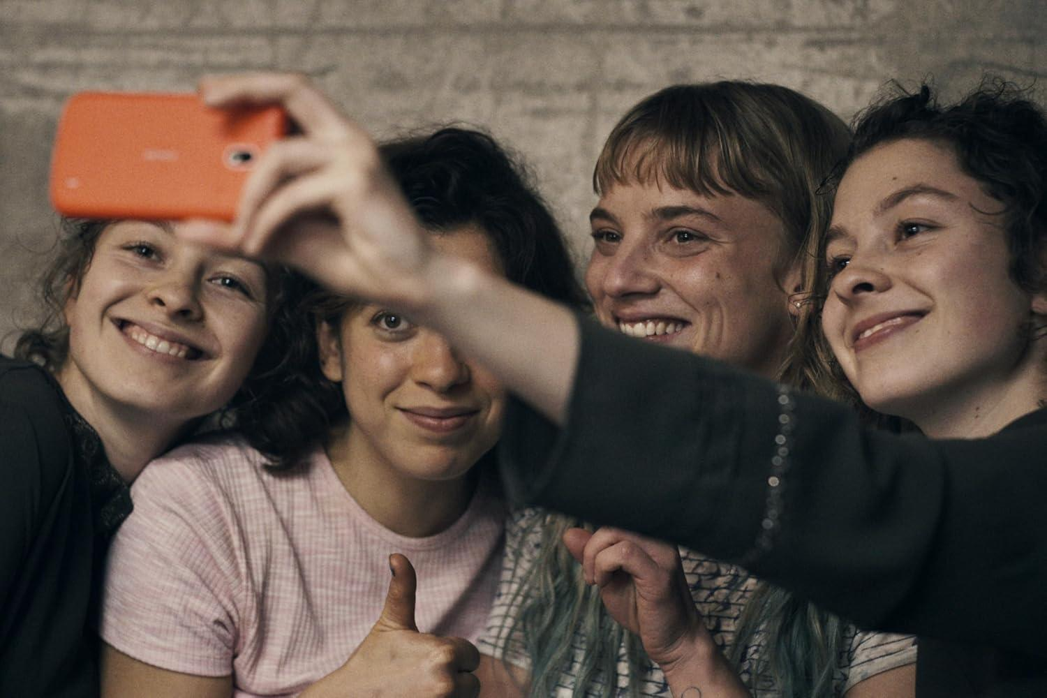 Vier junge Frauen machen ein Selfie mit einem orangefarbenen Handy. Sie lächeln.