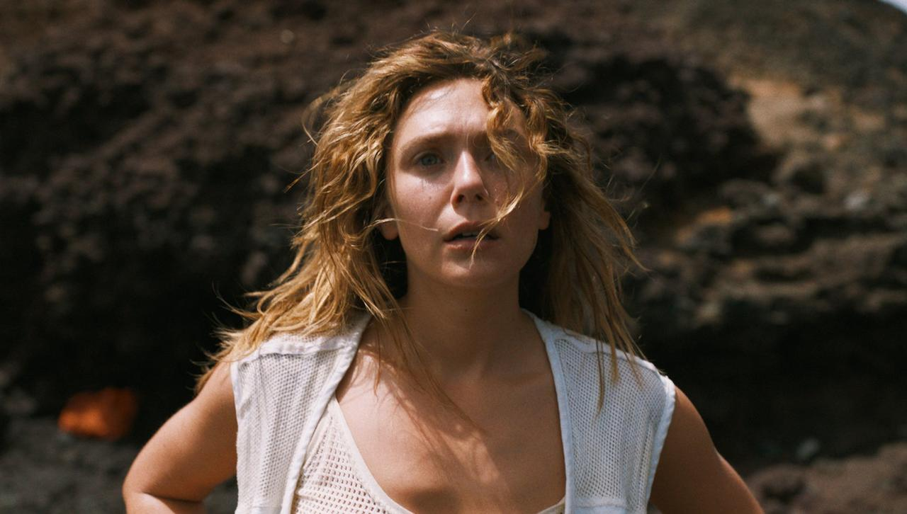 Eine Frau mit blonden Haaren steht vor einem Felsen und trägt ein weißes Oberteil.