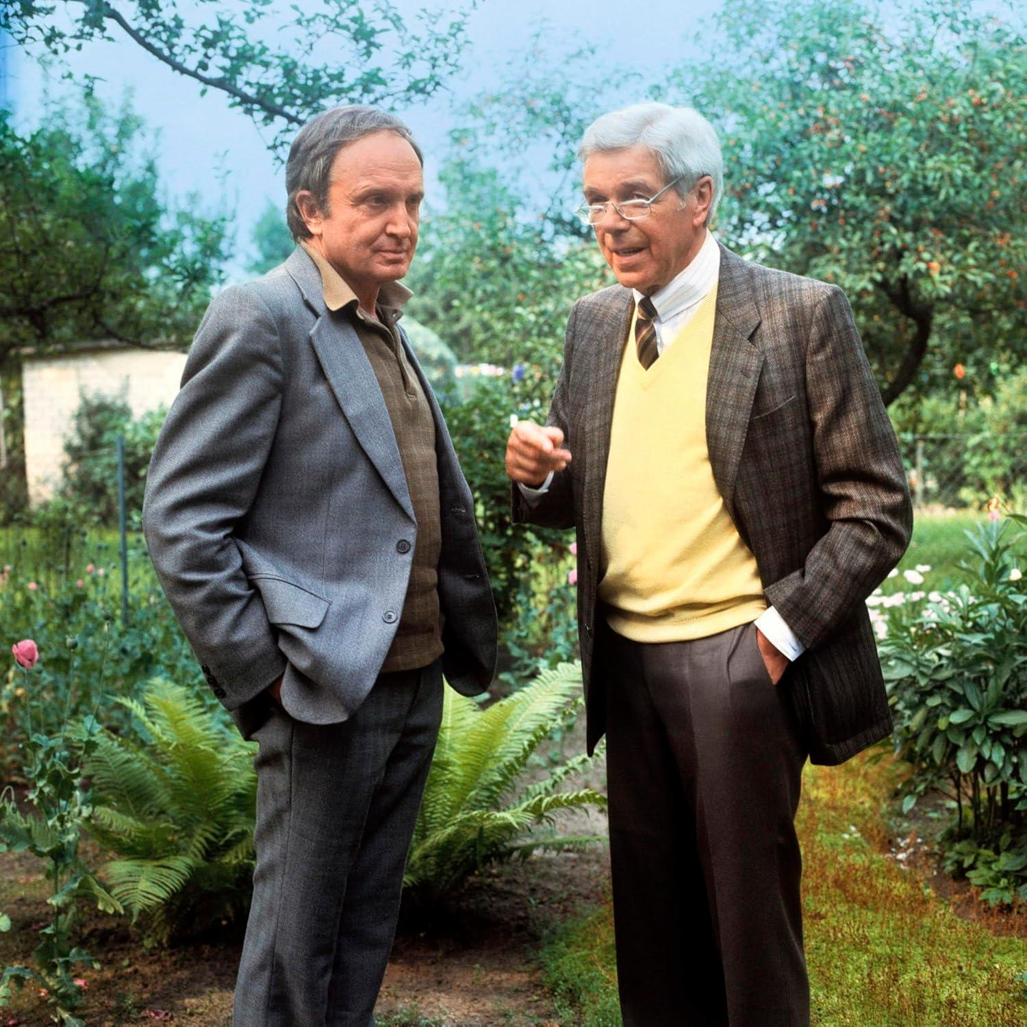 Zwei Männer stehen in einem Garten. Einer trägt einen grauen Anzug, der andere eine braune Jacke.