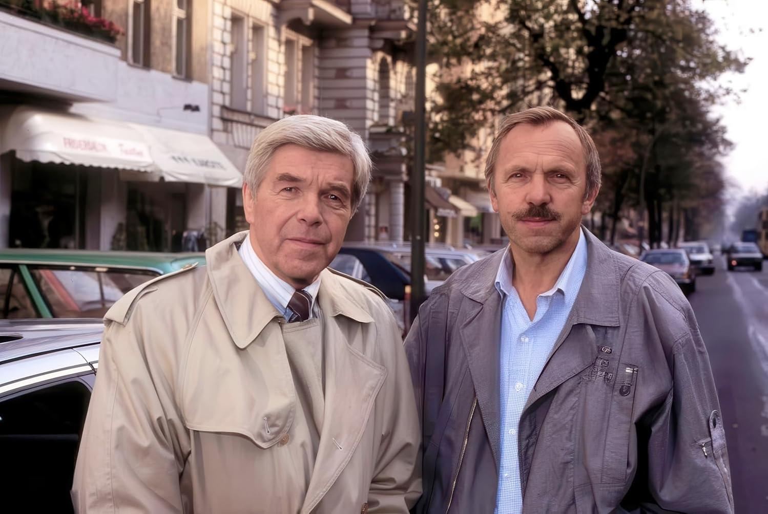 Zwei Männer stehen auf einer Straße. Einer trägt einen beigen Trenchcoat, der andere eine graue Jacke.