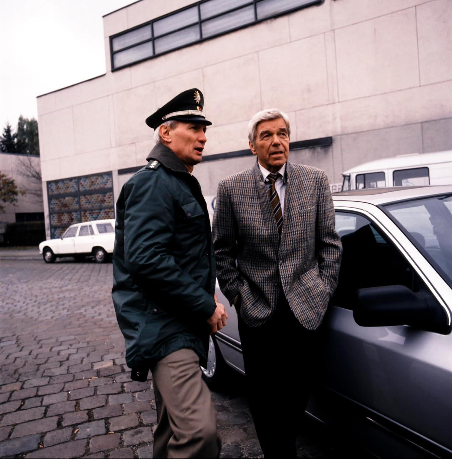 Zwei Männer in Anzügen stehen neben einem silbernen Auto. Einer trägt eine Uniform.