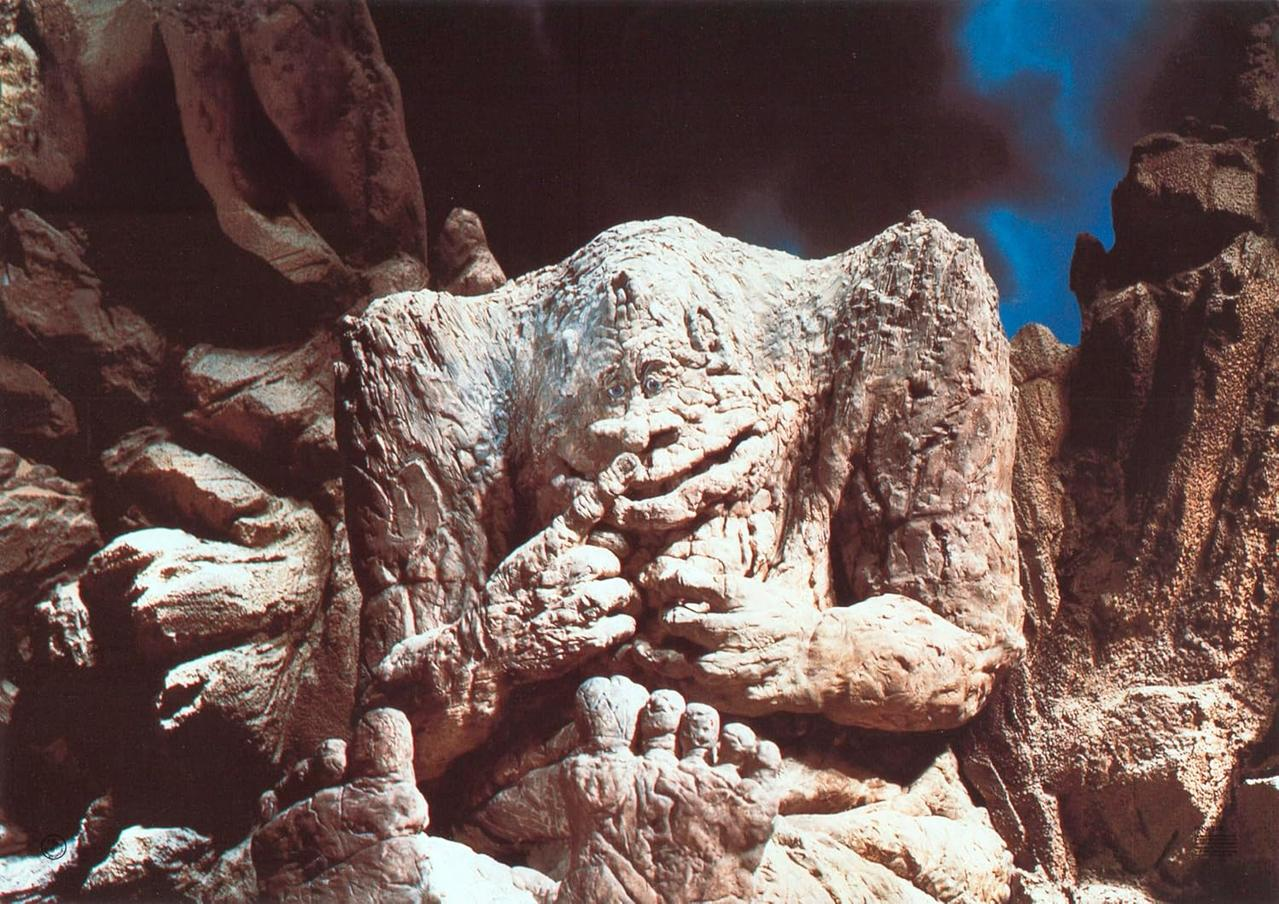 Ein Felsenwesen mit Gesicht und Händen, umgeben von Felsen und blauem Himmel.