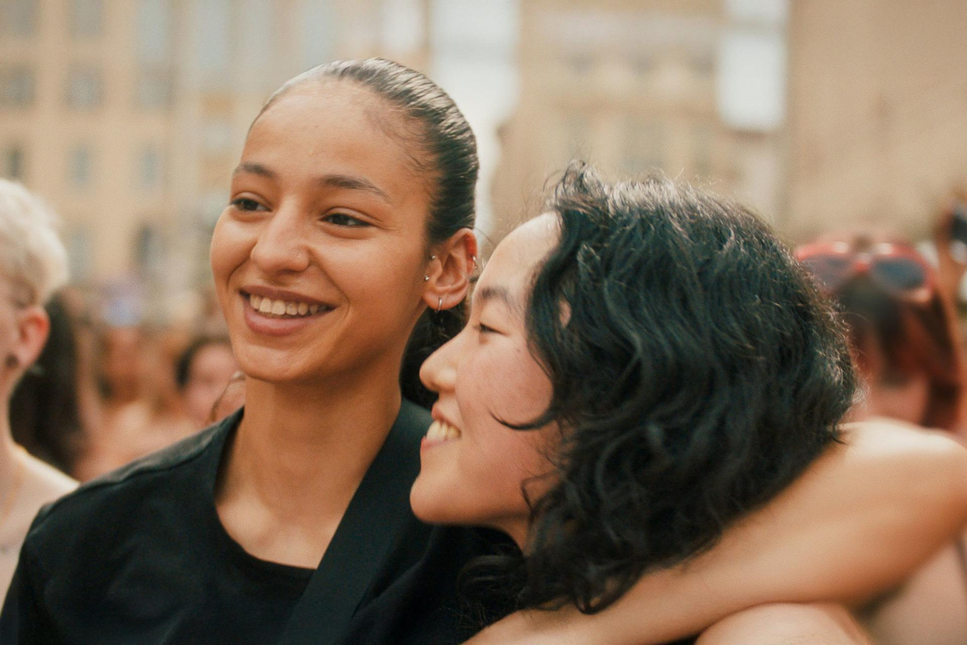 Zwei lächelnde Frauen umarmen sich in einer Menge. Die eine blickt nach links, die andere nach rechts.