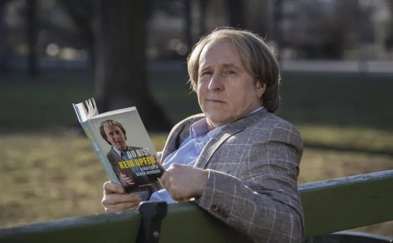 Ein Mann sitzt auf einer Parkbank und liest ein Buch mit seinem Bild auf dem Umschlag.