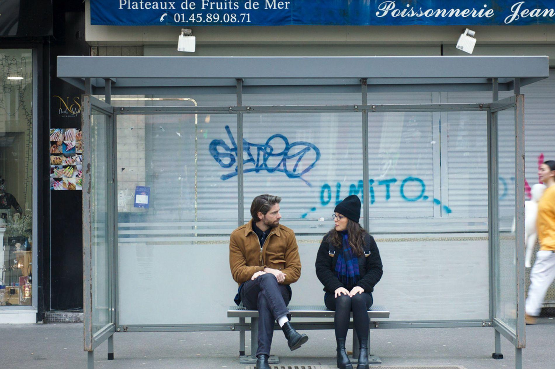Zwei Personen sitzen auf einer Bank an einer Bushaltestelle. Graffiti ist an der Wand.