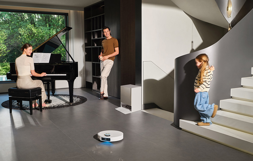 Odkurzacz automatyczny czyści podłogę, gdy kobieta gra na pianinie, a mężczyzna obserwuje.