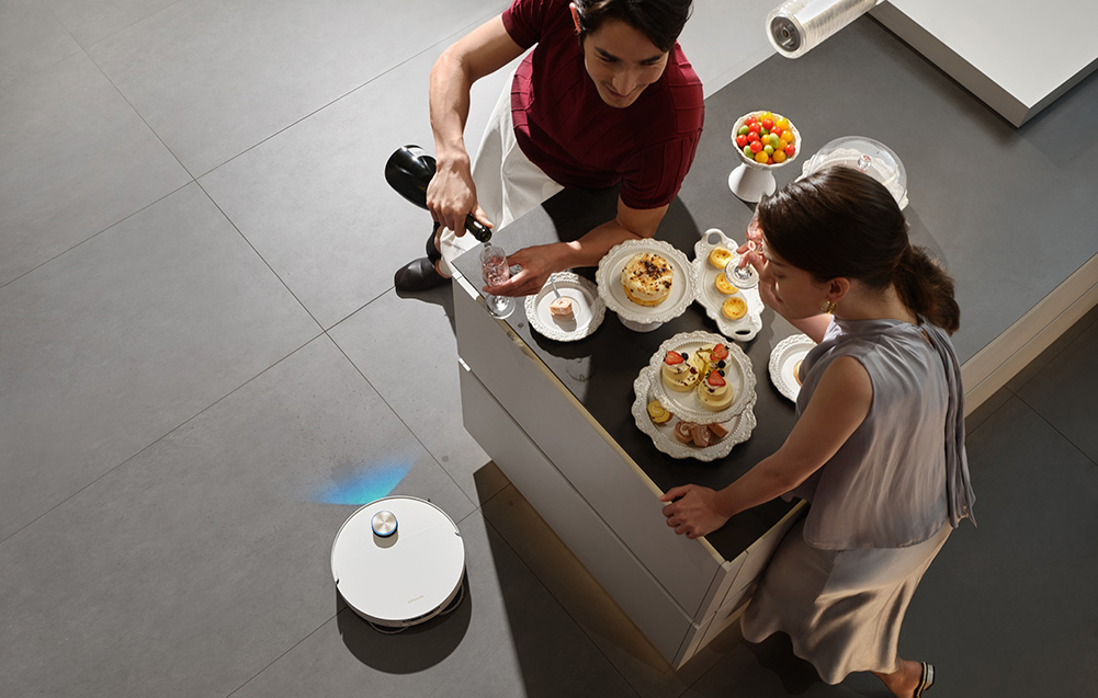 Mężczyzna nalewa szampana, podczas gdy robot sprzątający odkurza podłogę.