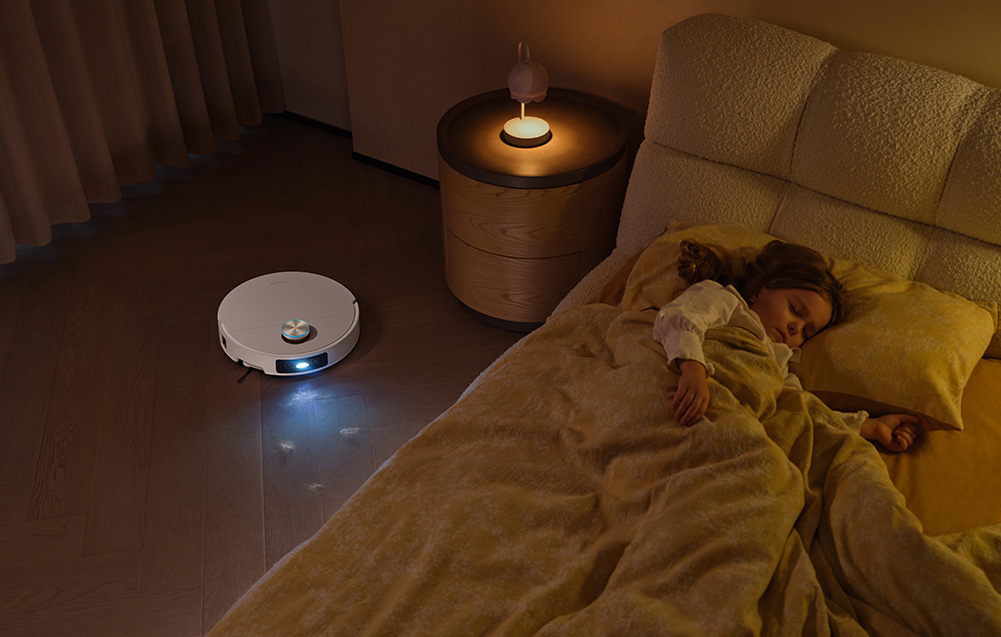 Robot sprzątający czyści drewnianą podłogę obok śpiącego dziecka w łóżku.