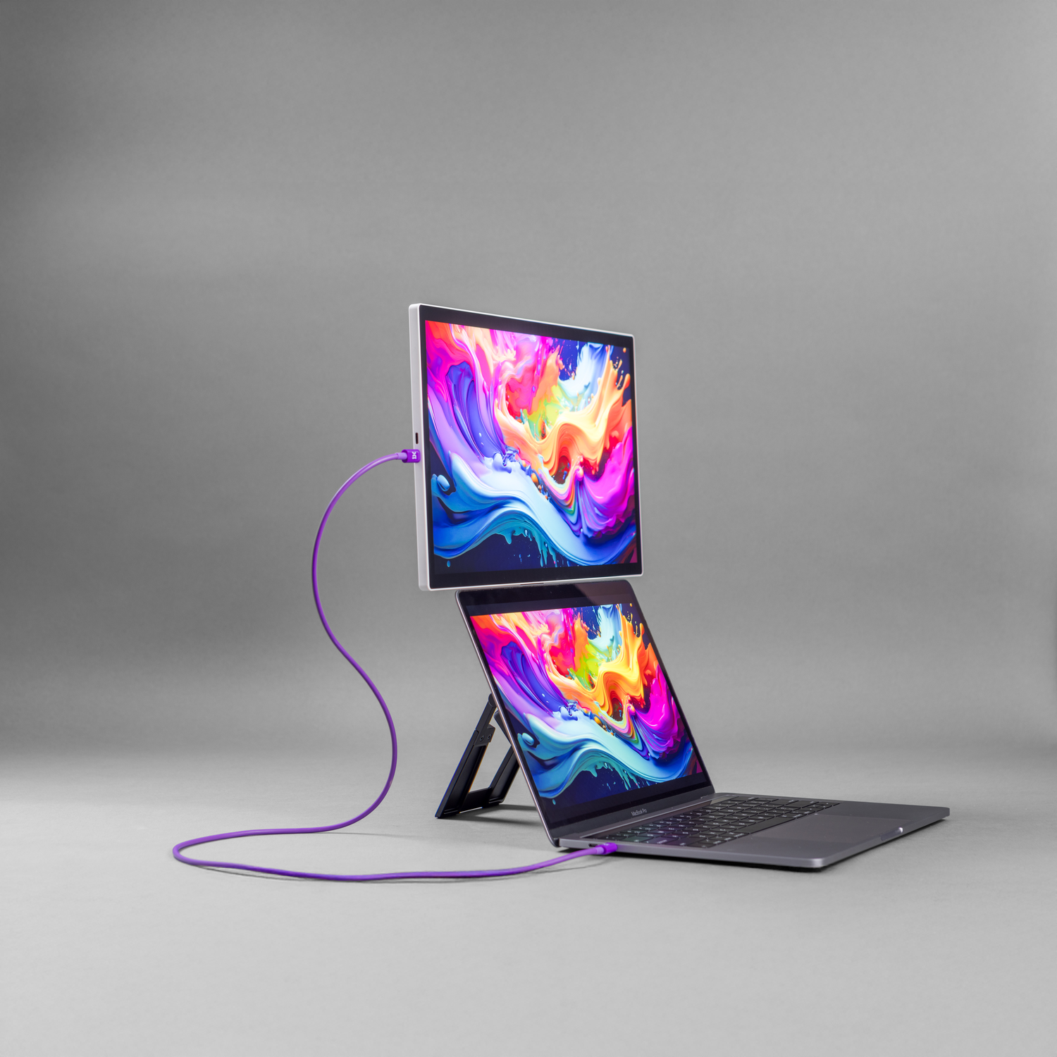 A laptop and external monitor stack, connected by a purple cable, display colorful abstract art.