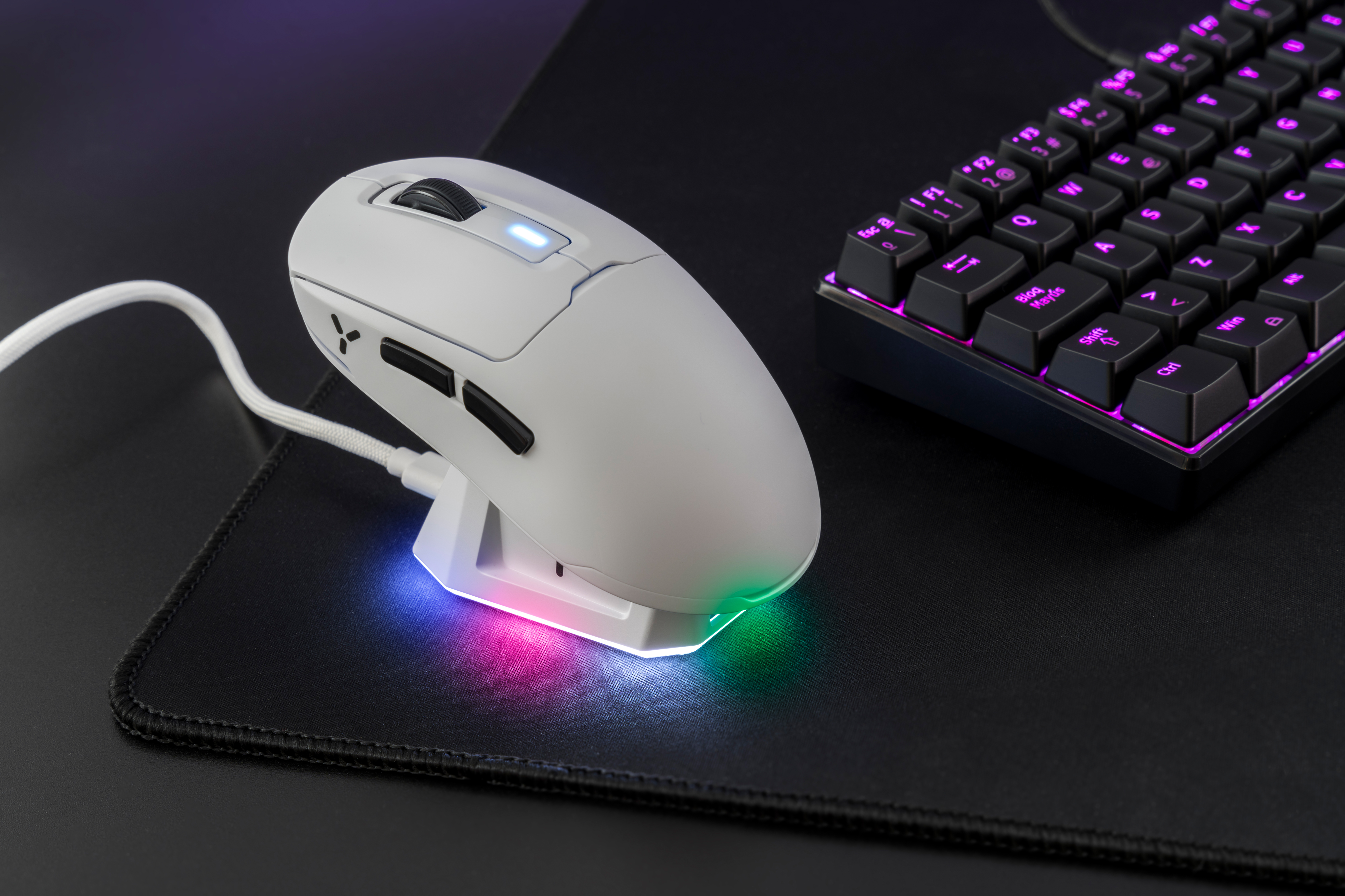 Biała mysz gamingowa z podświetleniem RGB leży na czarnej podkładce obok klawiatury.