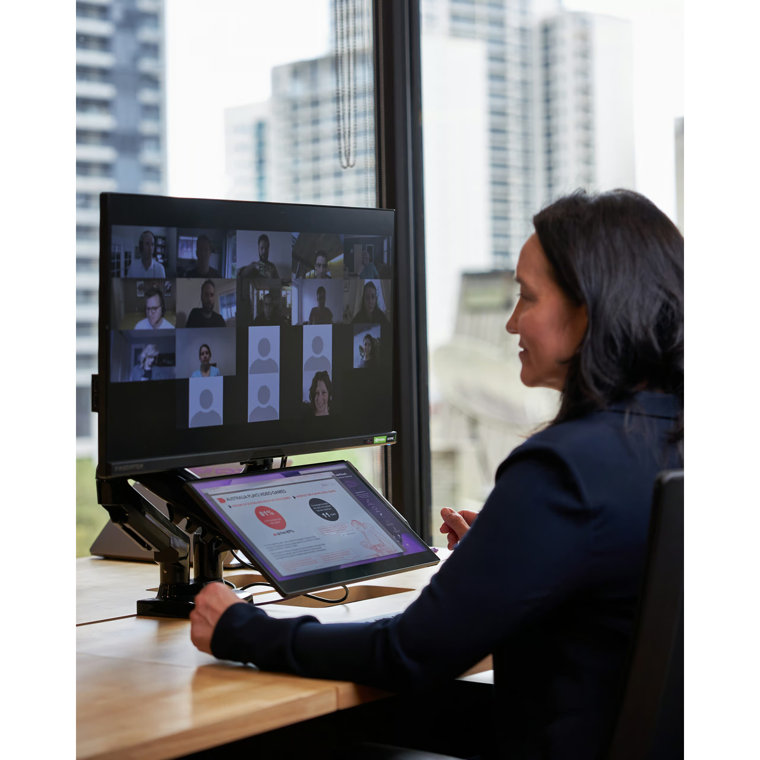 Woman in dark suit participates in video conference, monitors on desk. City view.