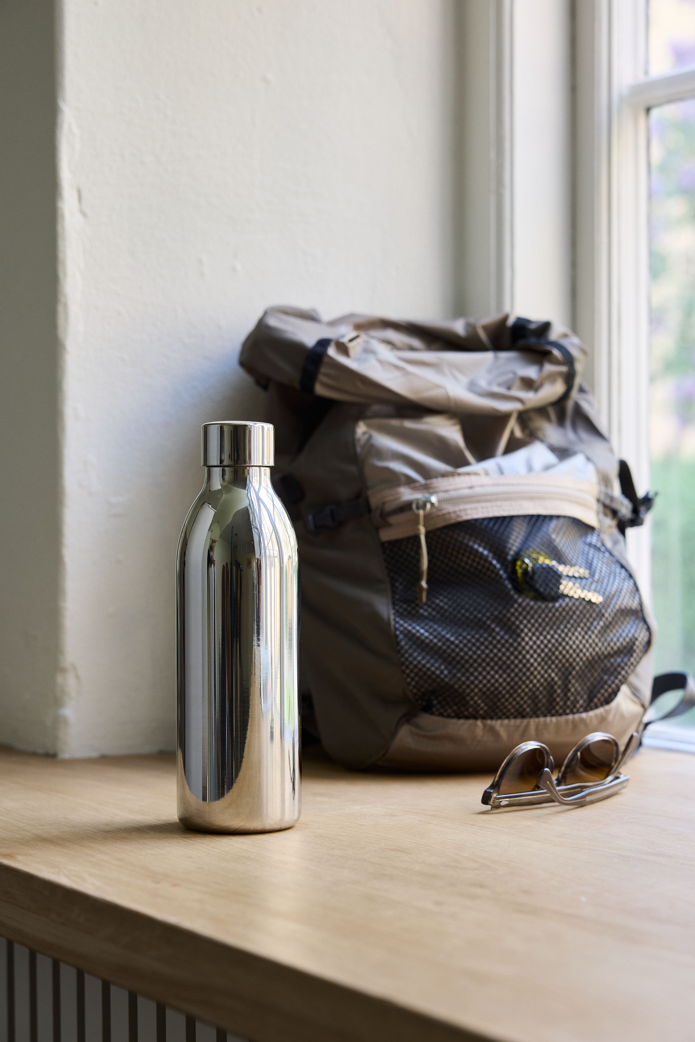 Eine silberne Flasche, ein Rucksack und eine Sonnenbrille auf einem Holztisch.