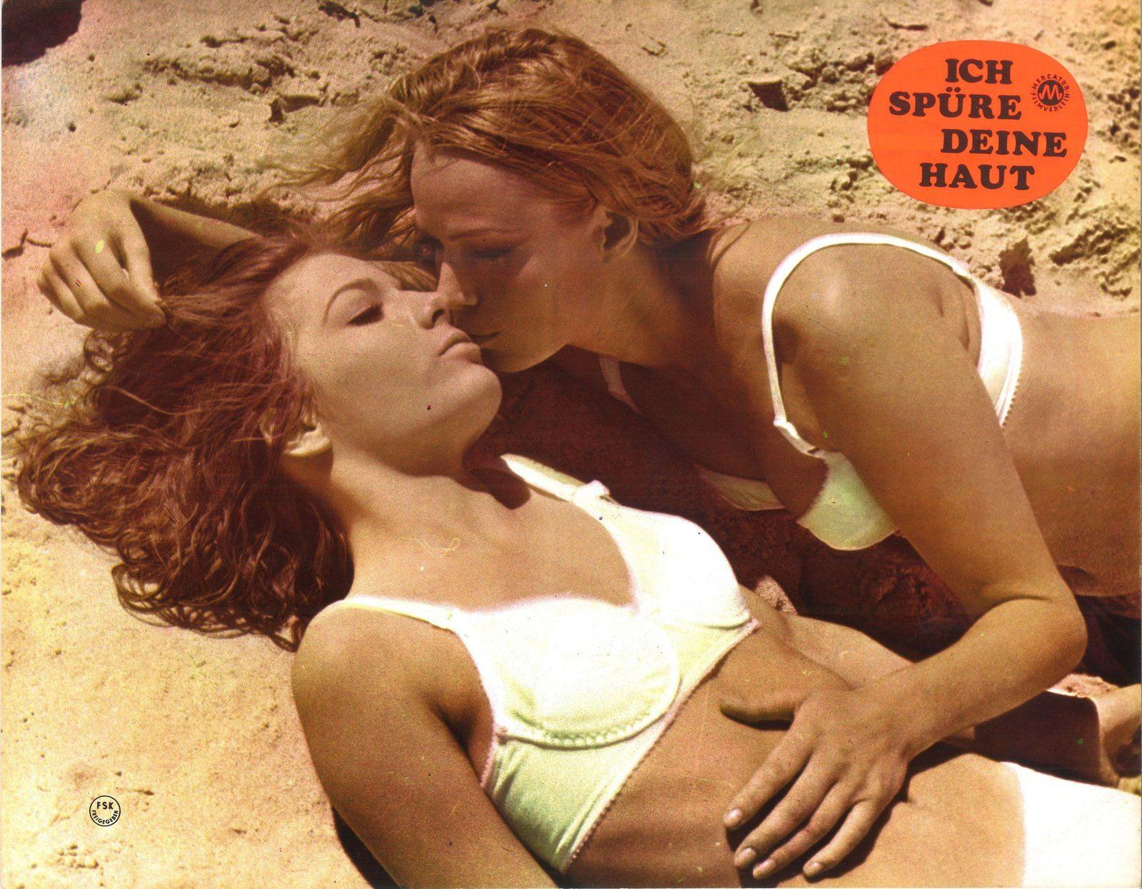Zwei Frauen in Bikinis liegen am Sandstrand, eine küsst die andere.