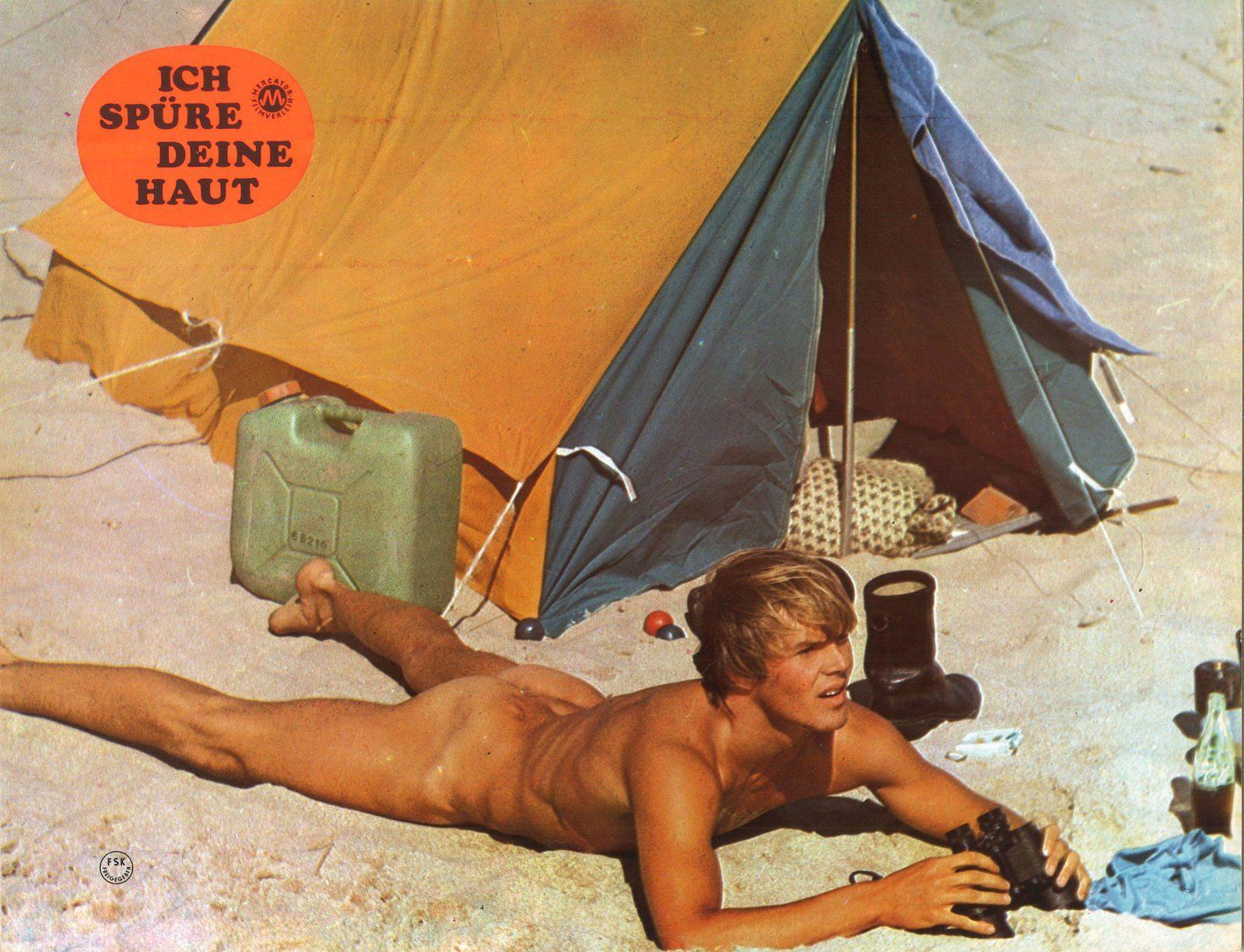 Ein Mann liegt auf dem Sand vor einem Zelt mit einem Kanister und Fernglas.