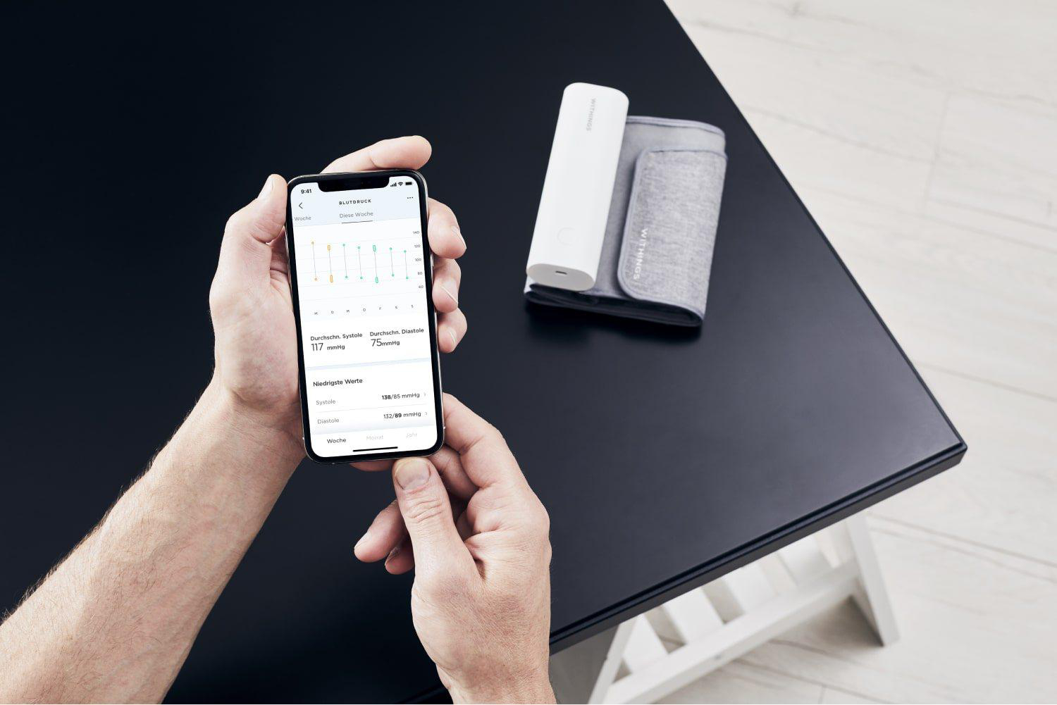 Eine Hand hält ein Smartphone mit einer Blutdruck-App. Daneben liegt ein Blutdruckmessgerät.