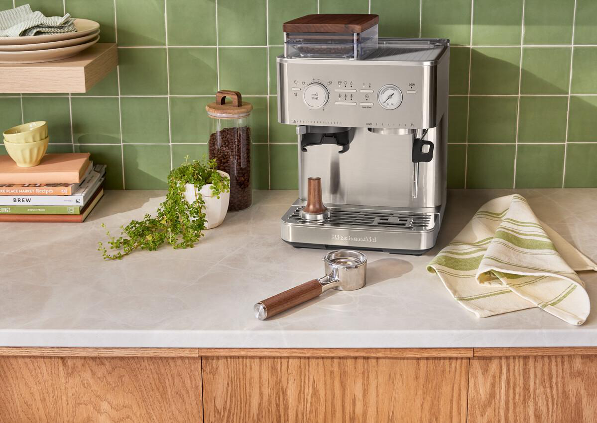 Silberne Kaffeemaschine mit Holzdetails neben Kaffeebohnenglas und grün gefliester Wand.