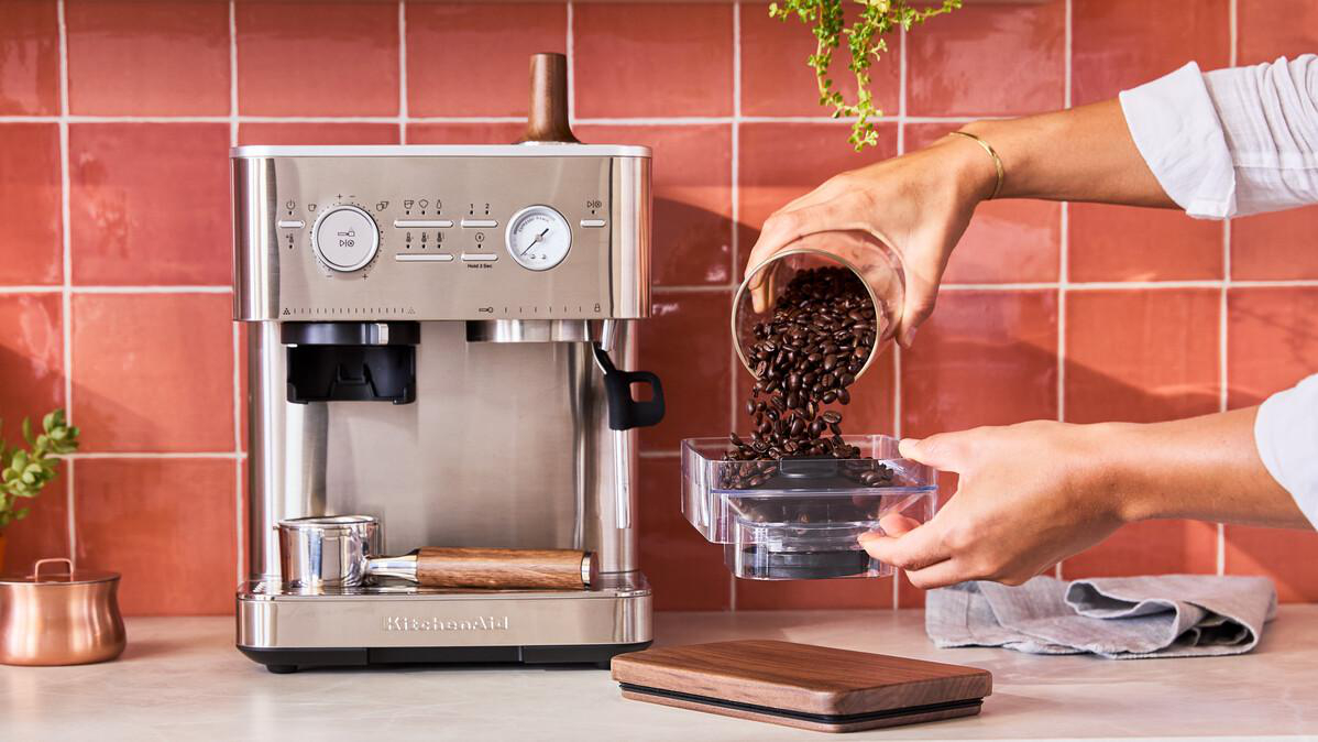 Eine Person gießt Kaffeebohnen aus einem Glas in einen Behälter neben einer Kaffeemaschine.