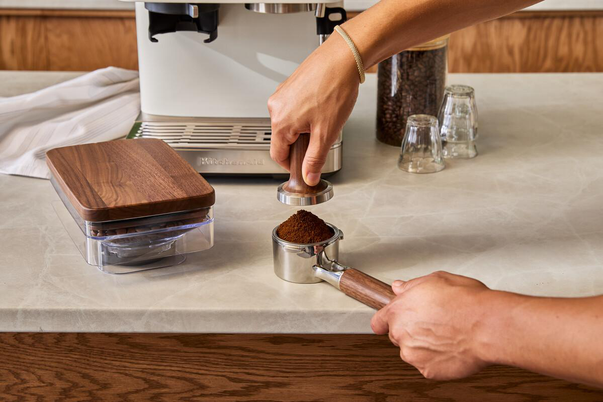 Eine Hand drückt Kaffeepulver mit einem Tamper in einen Siebträger.