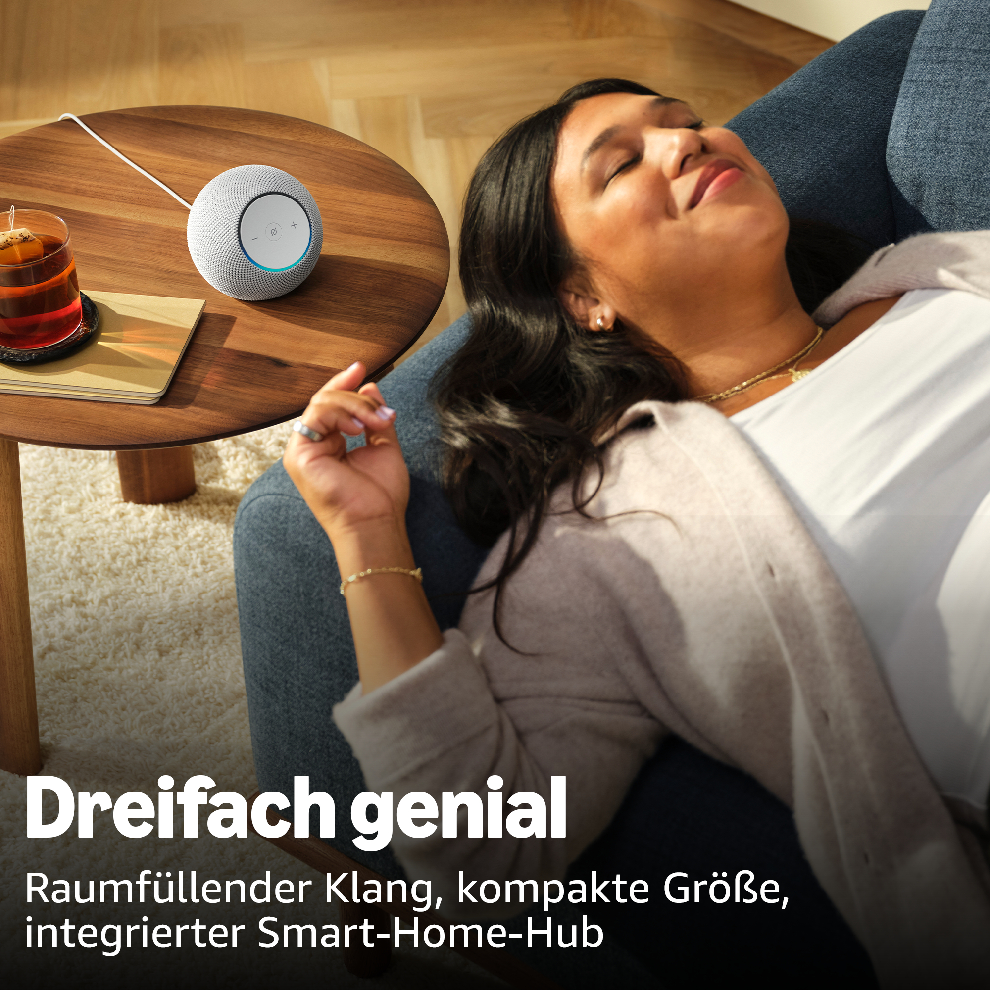 Eine Frau entspannt sich auf einem Sofa, neben ihr ein smarter Lautsprecher und ein Glas Tee.