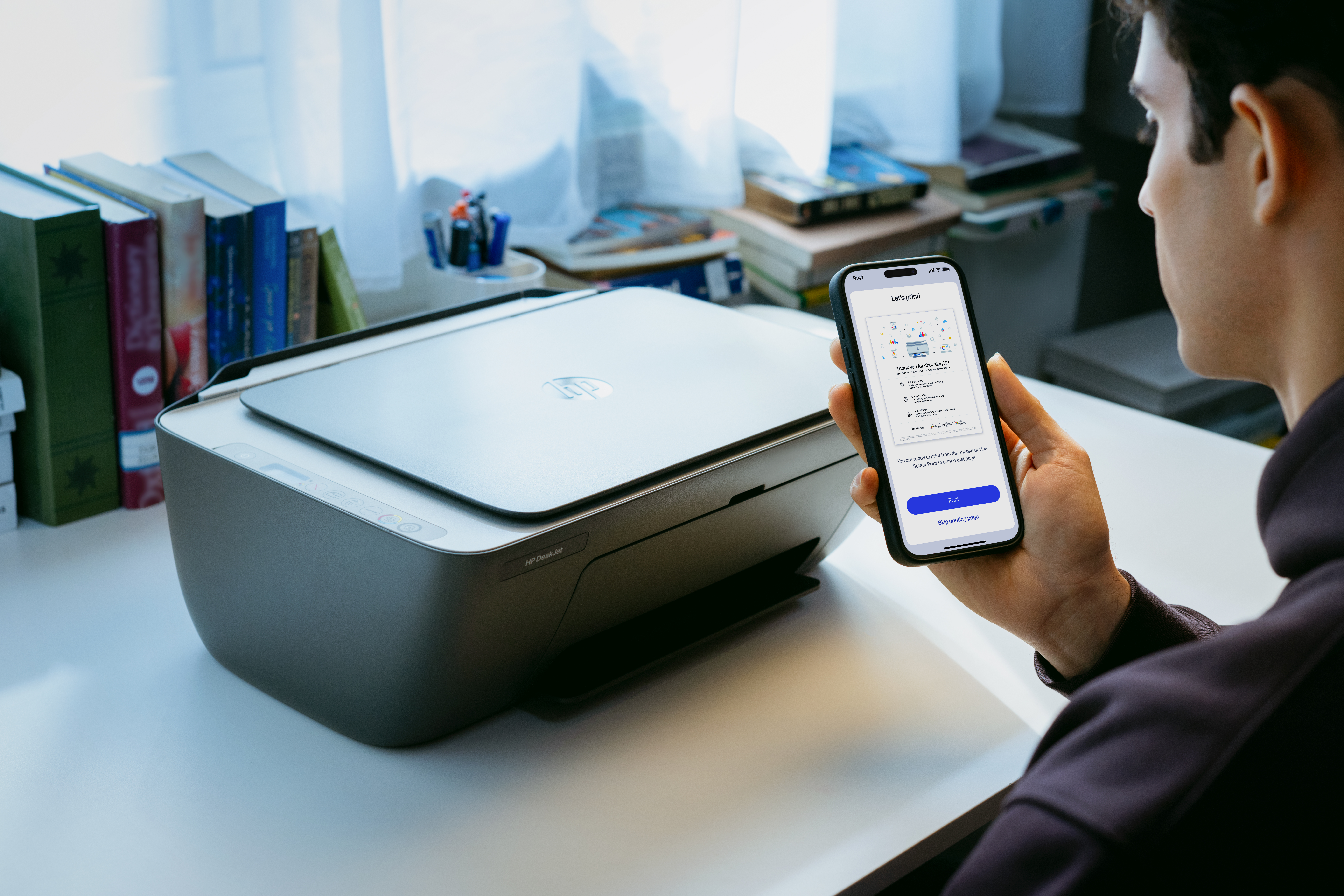 Ein Mann hält ein Smartphone vor einem Drucker. Bücher sind im Hintergrund.