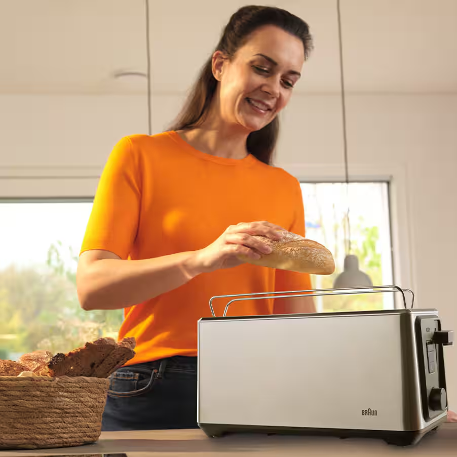 Femme en haut orange met une brioche dans un grille-pain argenté. Panier à pain à côté.