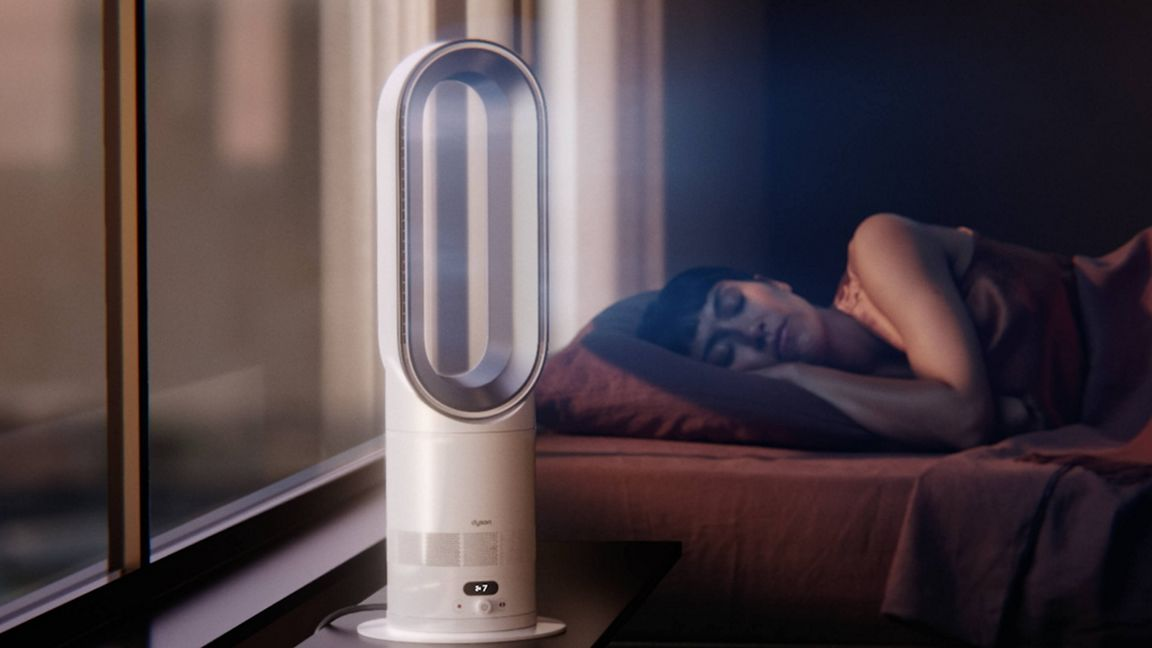 Un ventilateur Dyson blanc près d'une fenêtre, une femme dort dans un lit.
