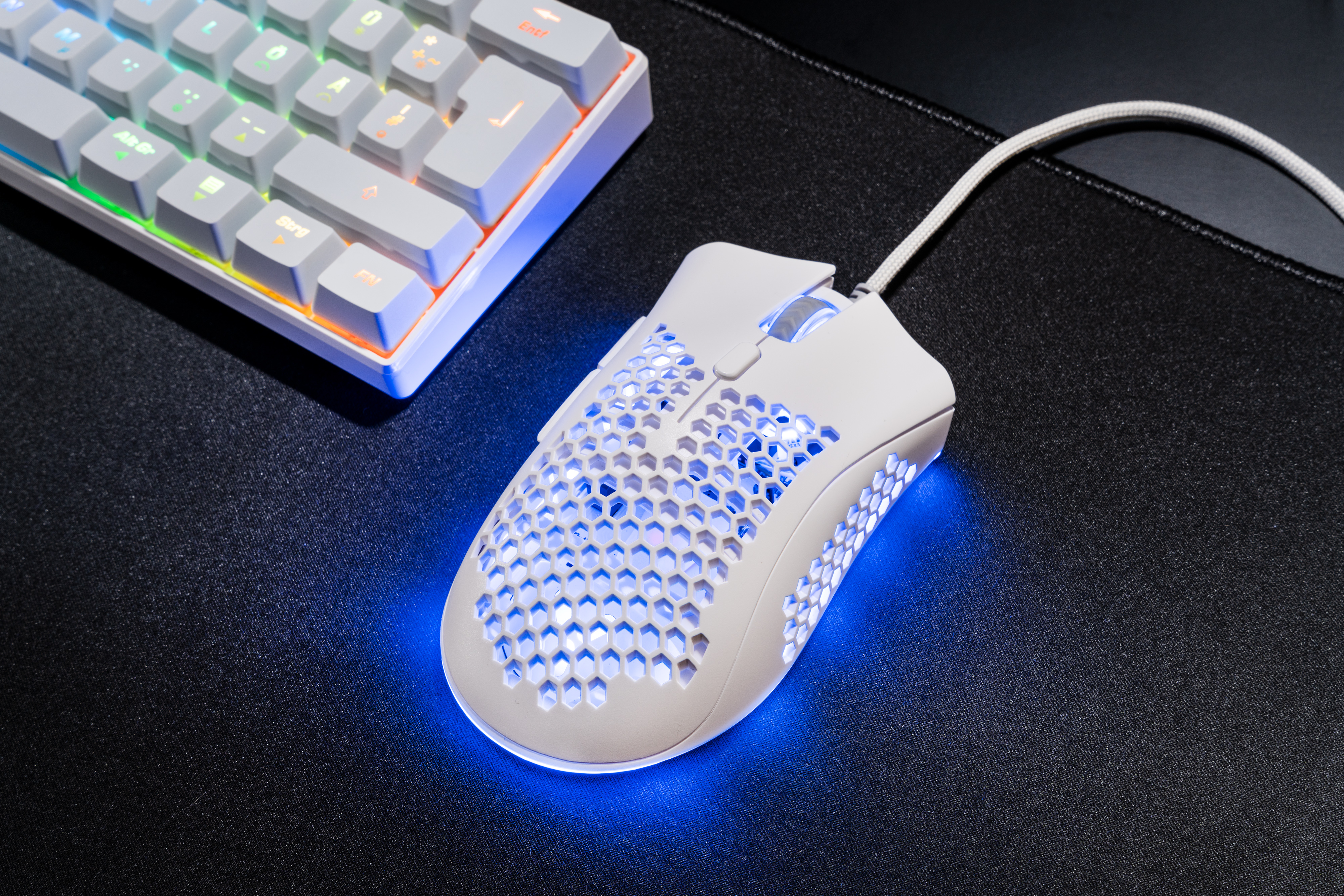 Biała mysz gamingowa z wzorem plastra miodu i niebieskim podświetleniem obok klawiatury.