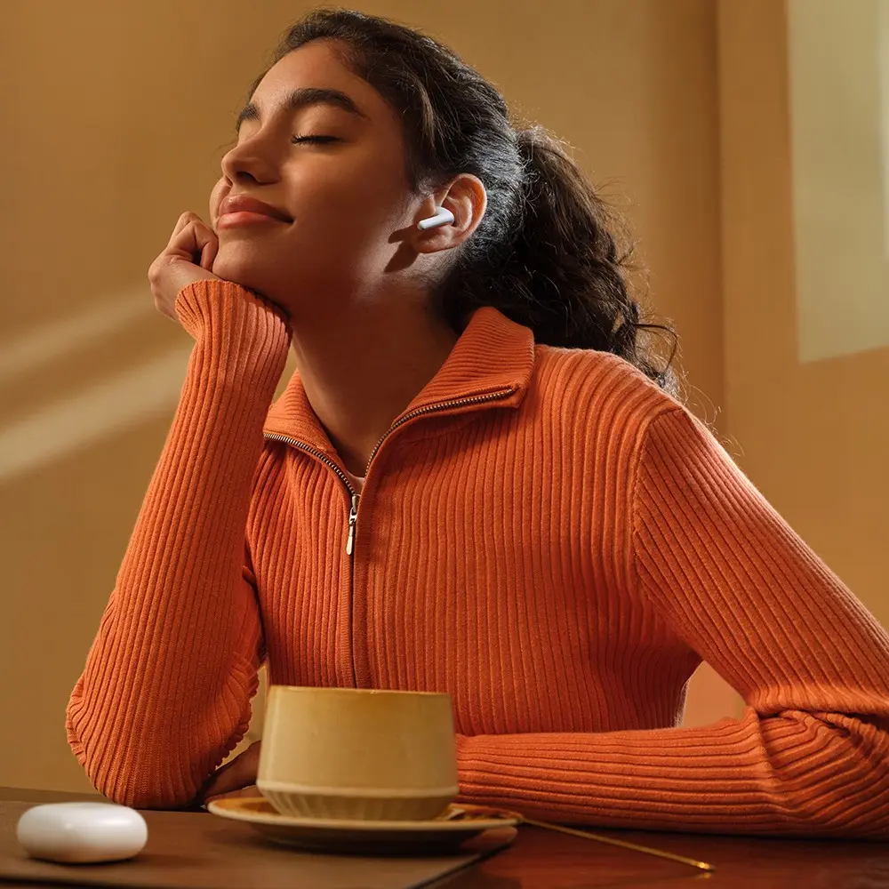 Eine junge Frau im orangen Pullover genießt Musik mit weißen Ohrhörern, Augen geschlossen.