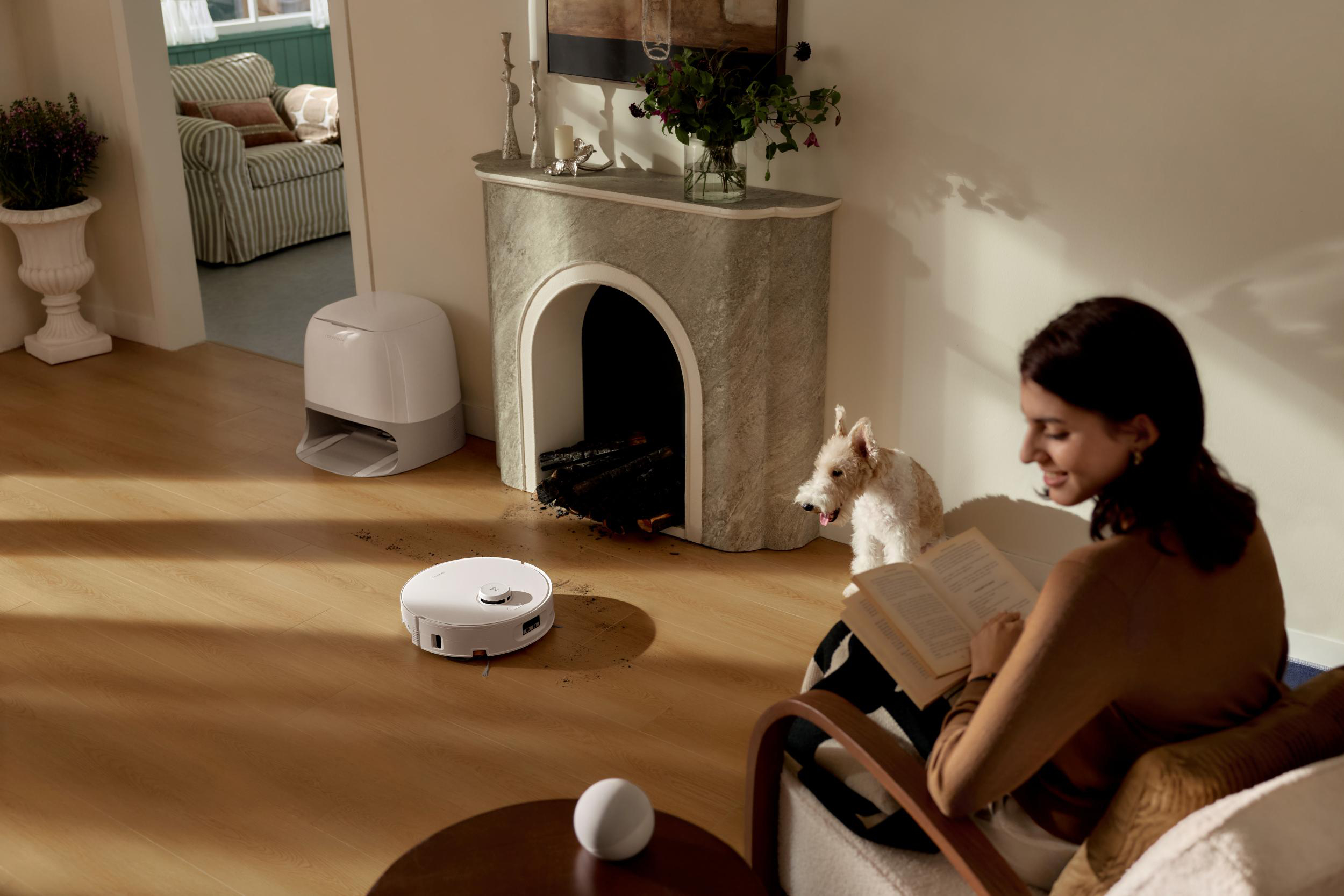 Ein Roboterstaubsauger reinigt einen Holzboden in der Nähe eines Kamins, während ein Hund und eine Frau lesen.