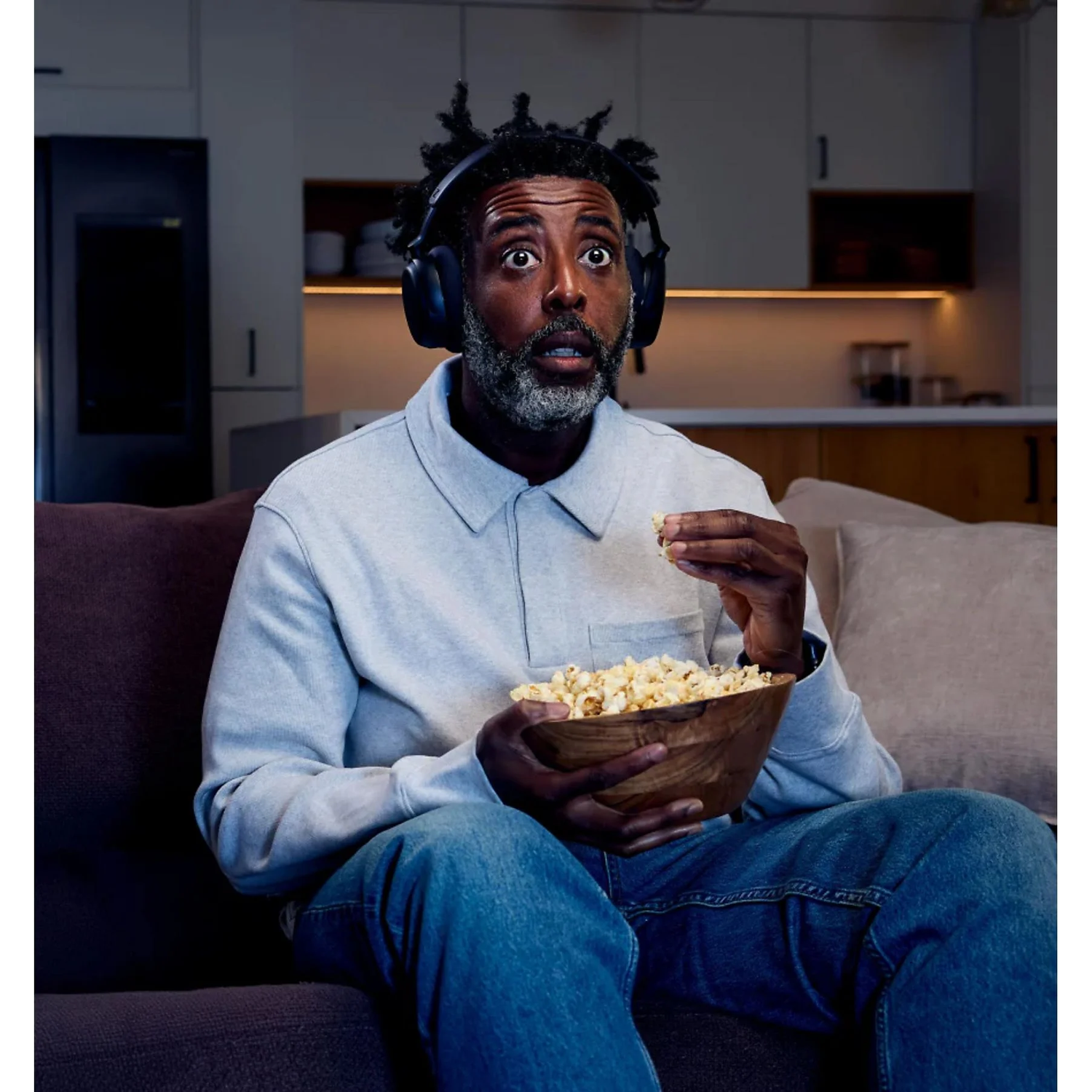 Man wearing headphones eats popcorn while sitting on a couch.