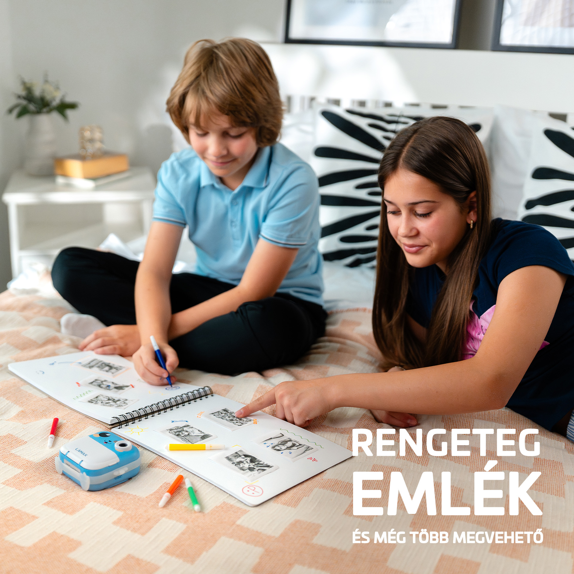 Két gyerek ül egy ágyon, vázlatfüzetbe rajzolnak. Egy kék-fehér fényképezőgép van mellettük.