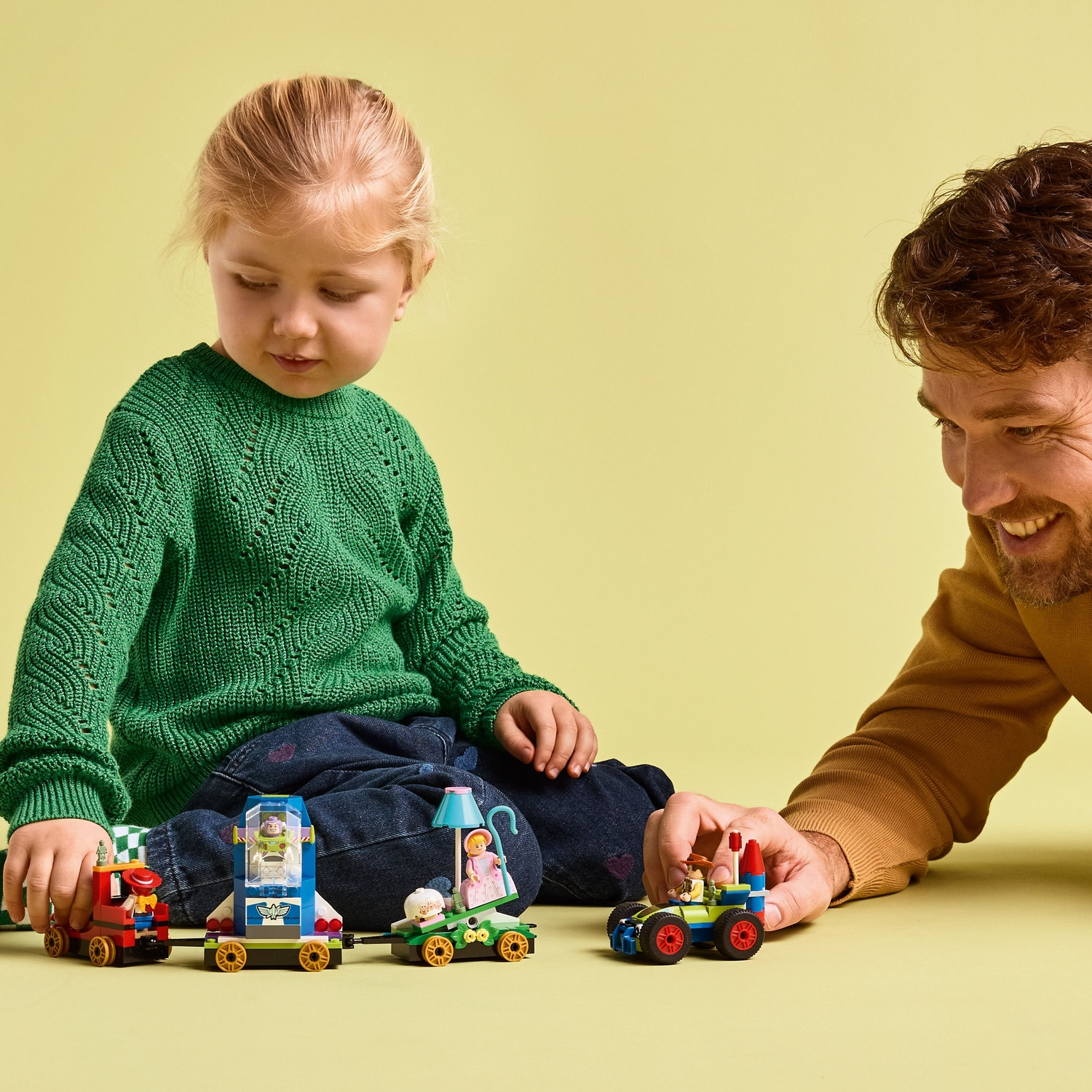 Père et fille jouent avec un train et une voiture LEGO colorés sur fond jaune.