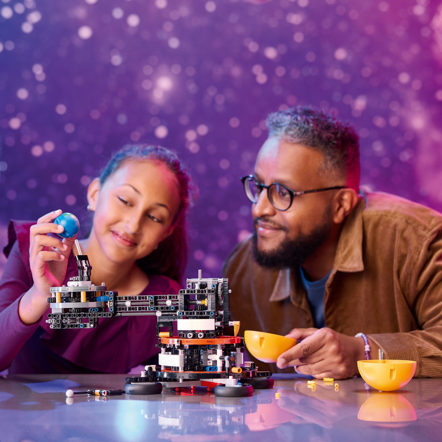 A girl holds a blue ball over a Lego build, a man beside her.