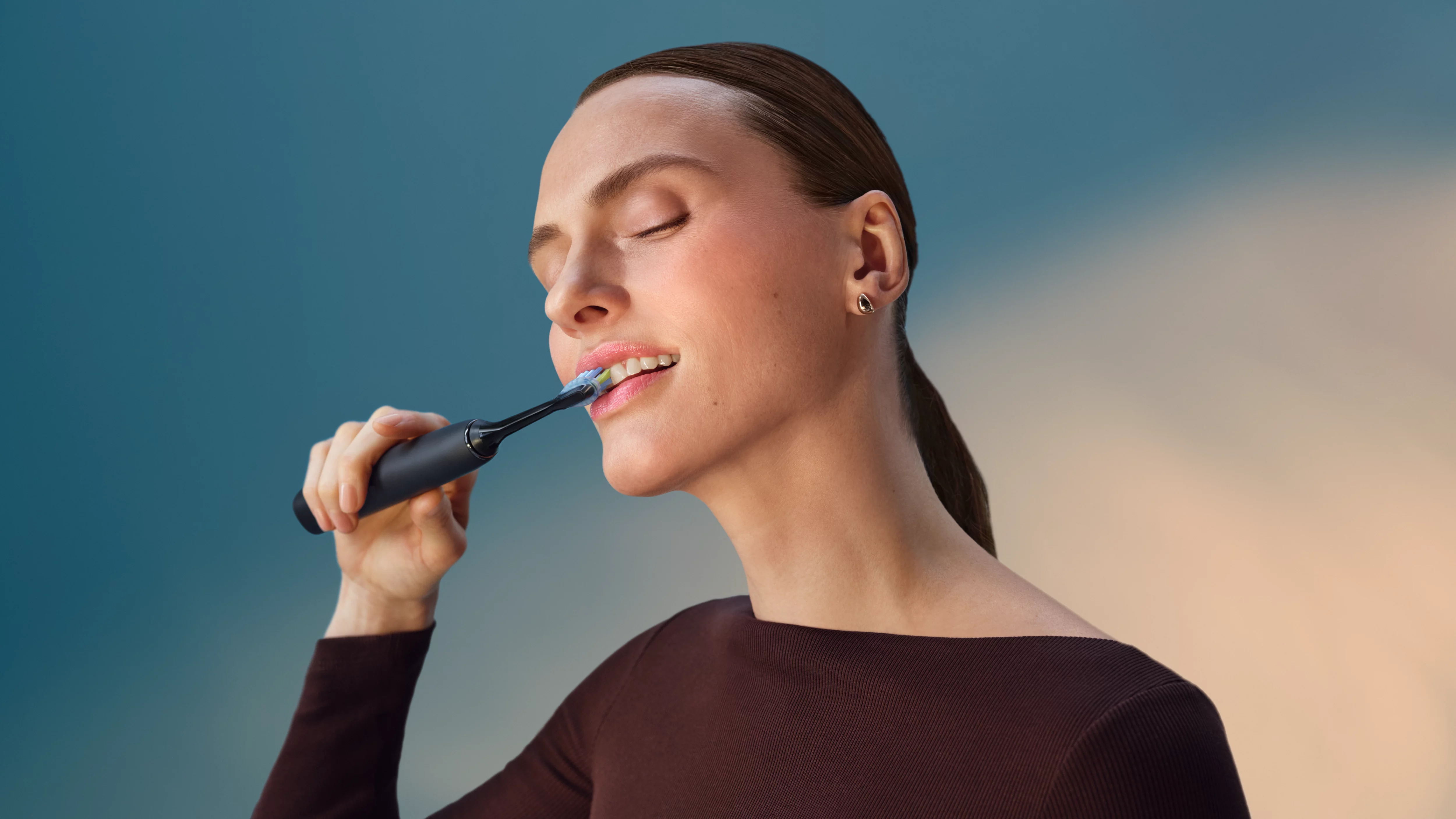 Kobieta myje zęby szczoteczką elektryczną na tle gradientu niebieskiego i pomarańczowego.