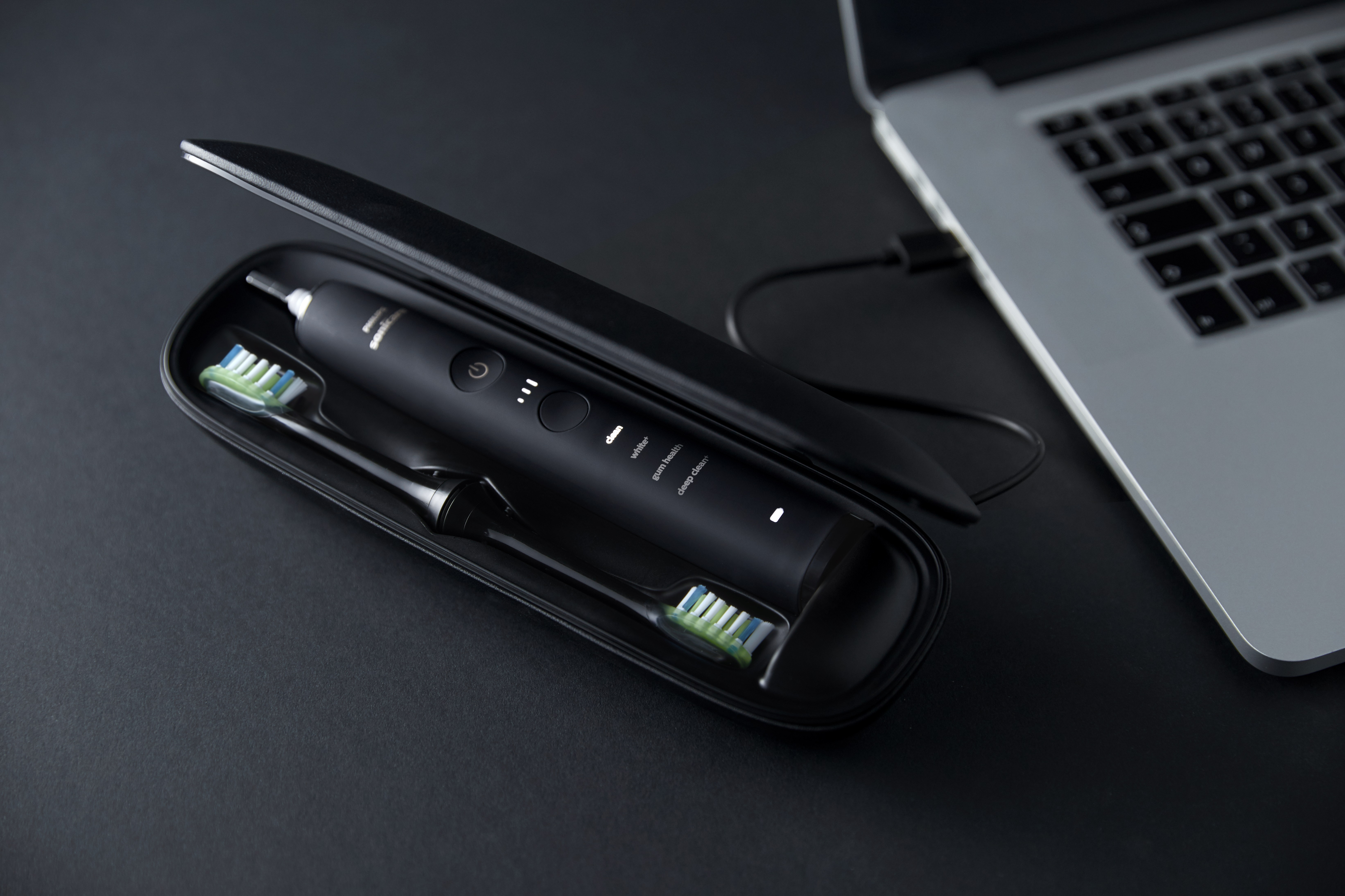 Czarna szczoteczka elektryczna z dwoma końcówkami w etui ładującym obok laptopa.